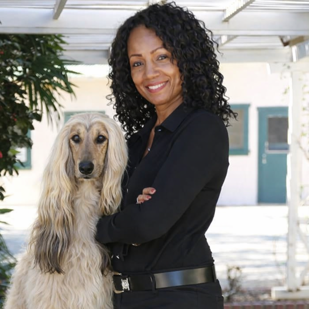 A woman with curly black hair smiling and holding a long-haired dog outside near a house.