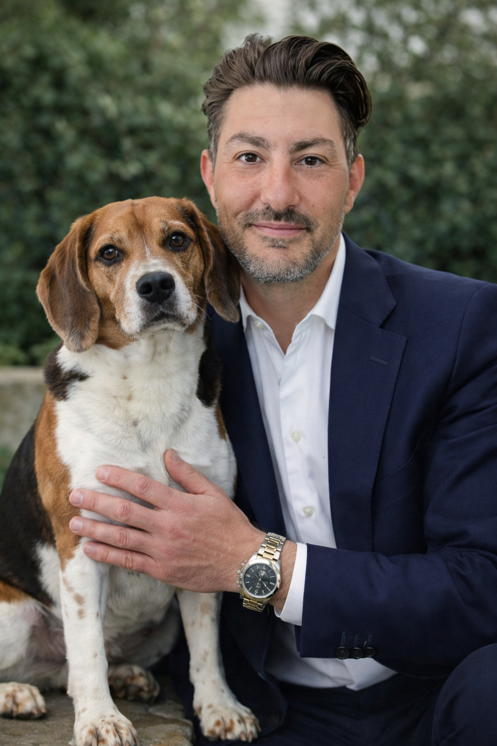 Man in a blue suit and white shirt holding a beagle dog outdoors with greenery in the background.