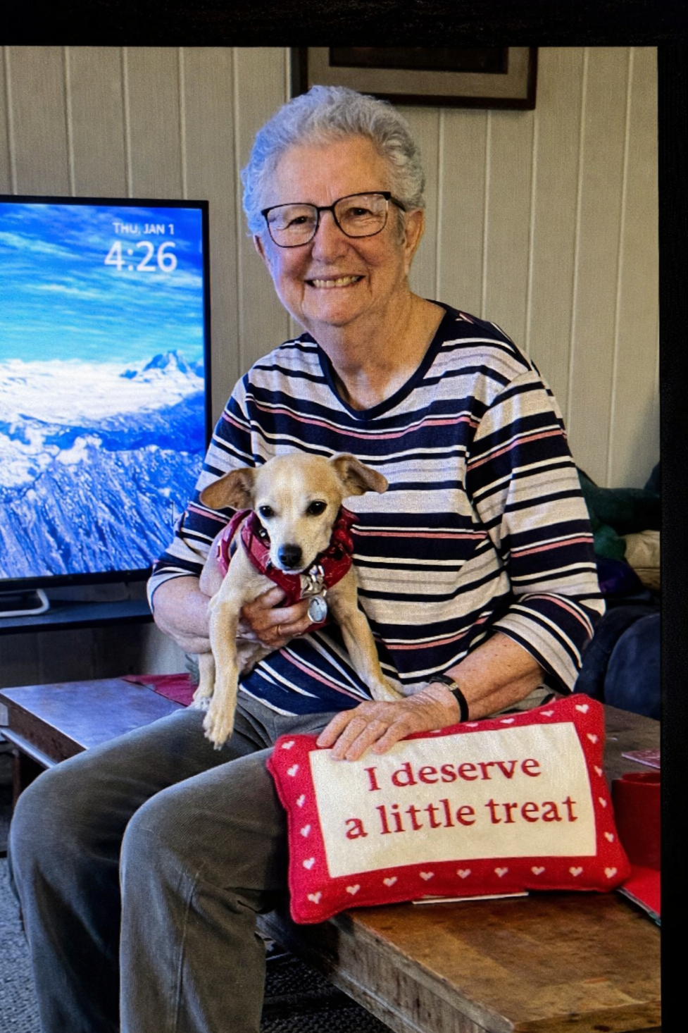 An elderly woman with gray hair and glasses smiling, holding a small dog wearing a red harness, sitting on a wooden bench with a cushion that reads "I deserve a little treat." In the background, there is a television screen showing mountains and a blue sky, with the time 4:26 and the date Thursday, January 1.