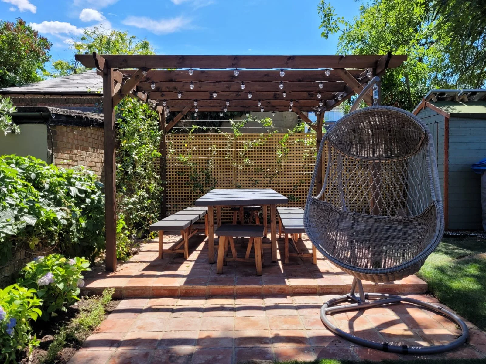 A backyard patio with a wooden pergola decorated with string lights, a picnic table with benches, and a hanging egg-shaped wicker chair, surrounded by greenery and a blue sky.