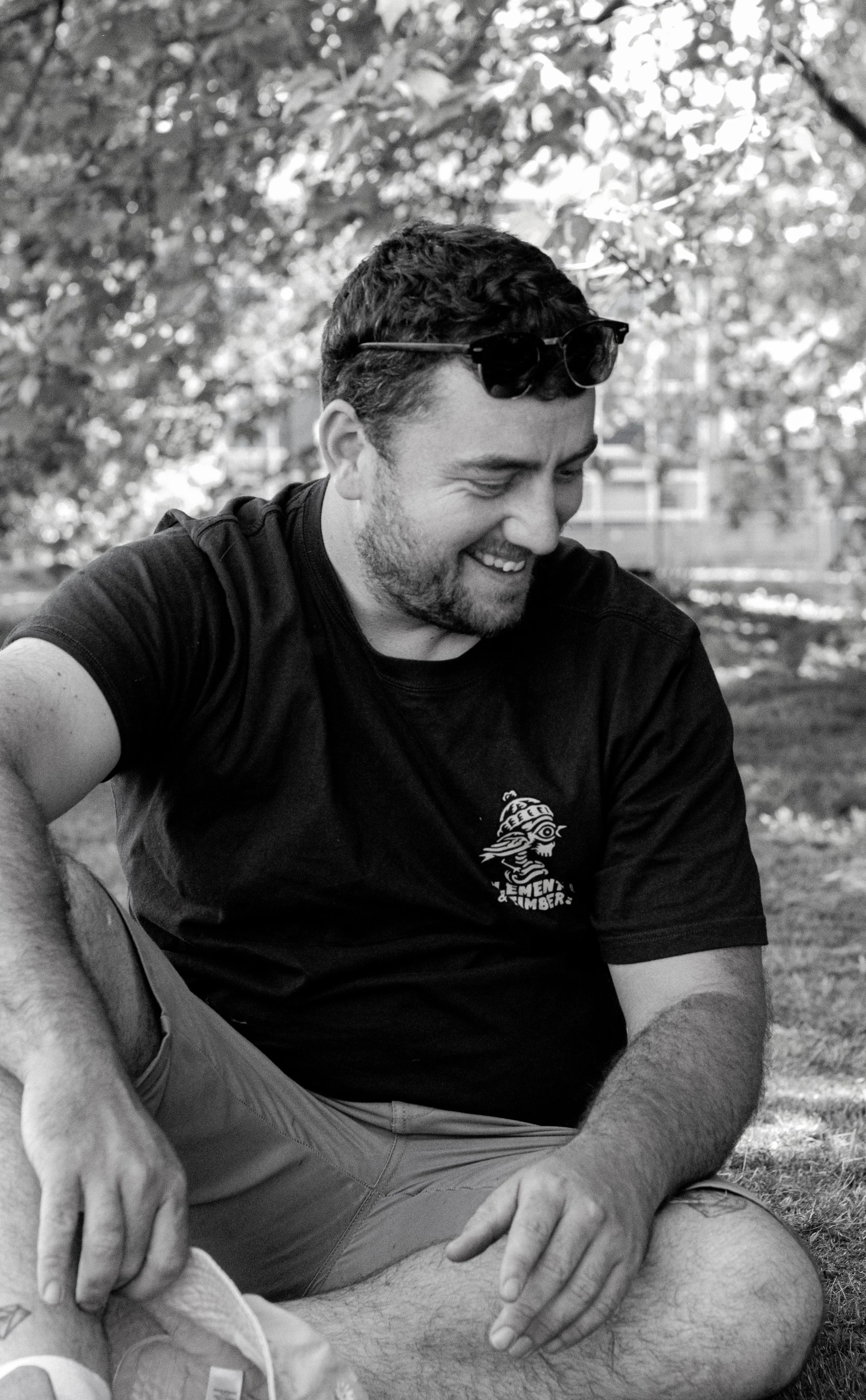 A smiling man sitting outdoors on the grass, with trees in the background, wearing a black t-shirt and sunglasses on his head.