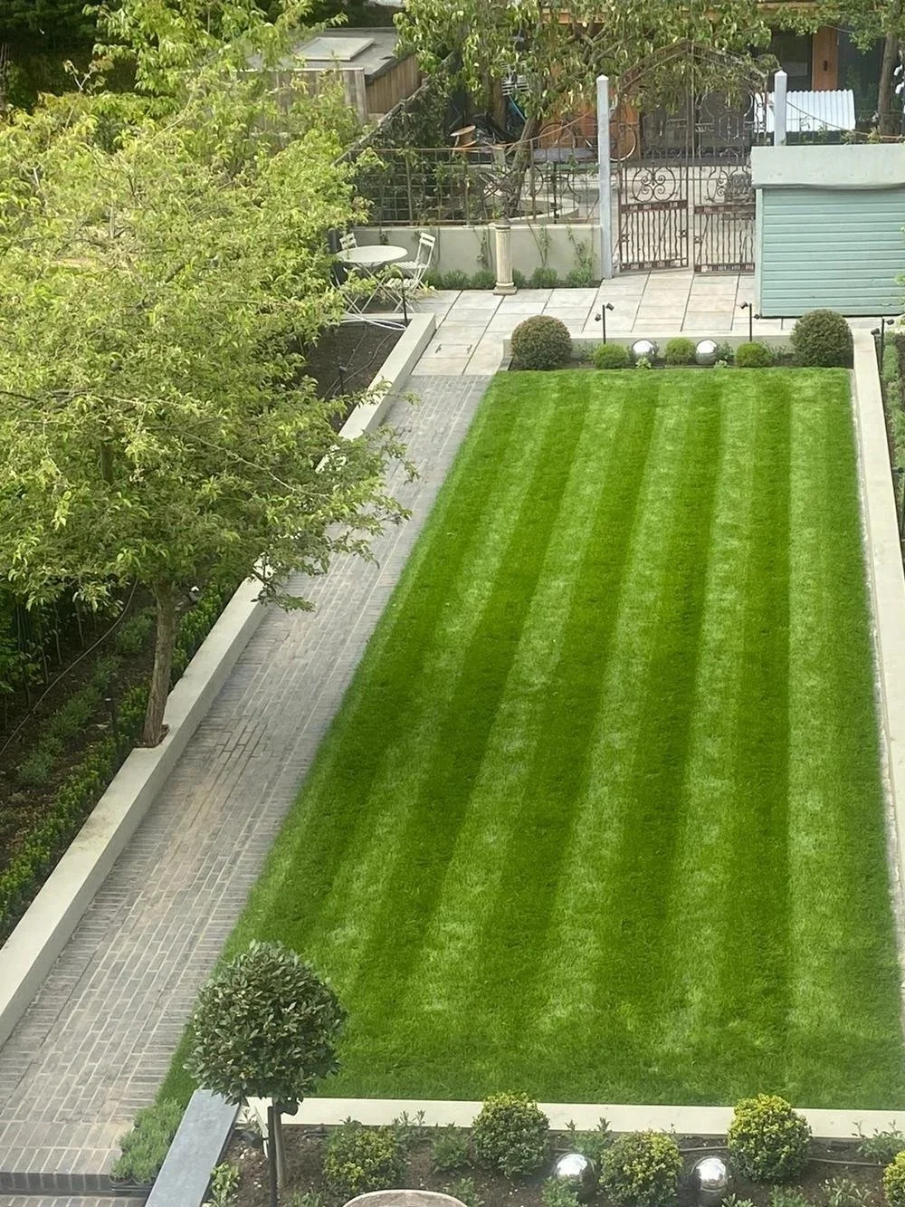 A backyard with a neatly mowed green lawn, bordered by small bushes and gravel pathway, with trees and garden furniture in the background.