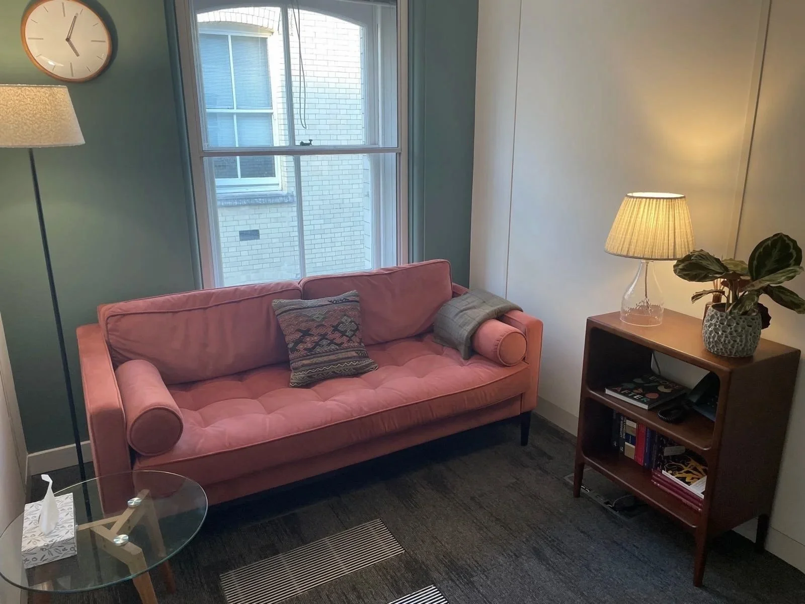 A cozy therapist' room with a velvet sofa, a patterned cushion, a gray blanket, a wooden side table with a glass lamp, a potted plant, and a window.
