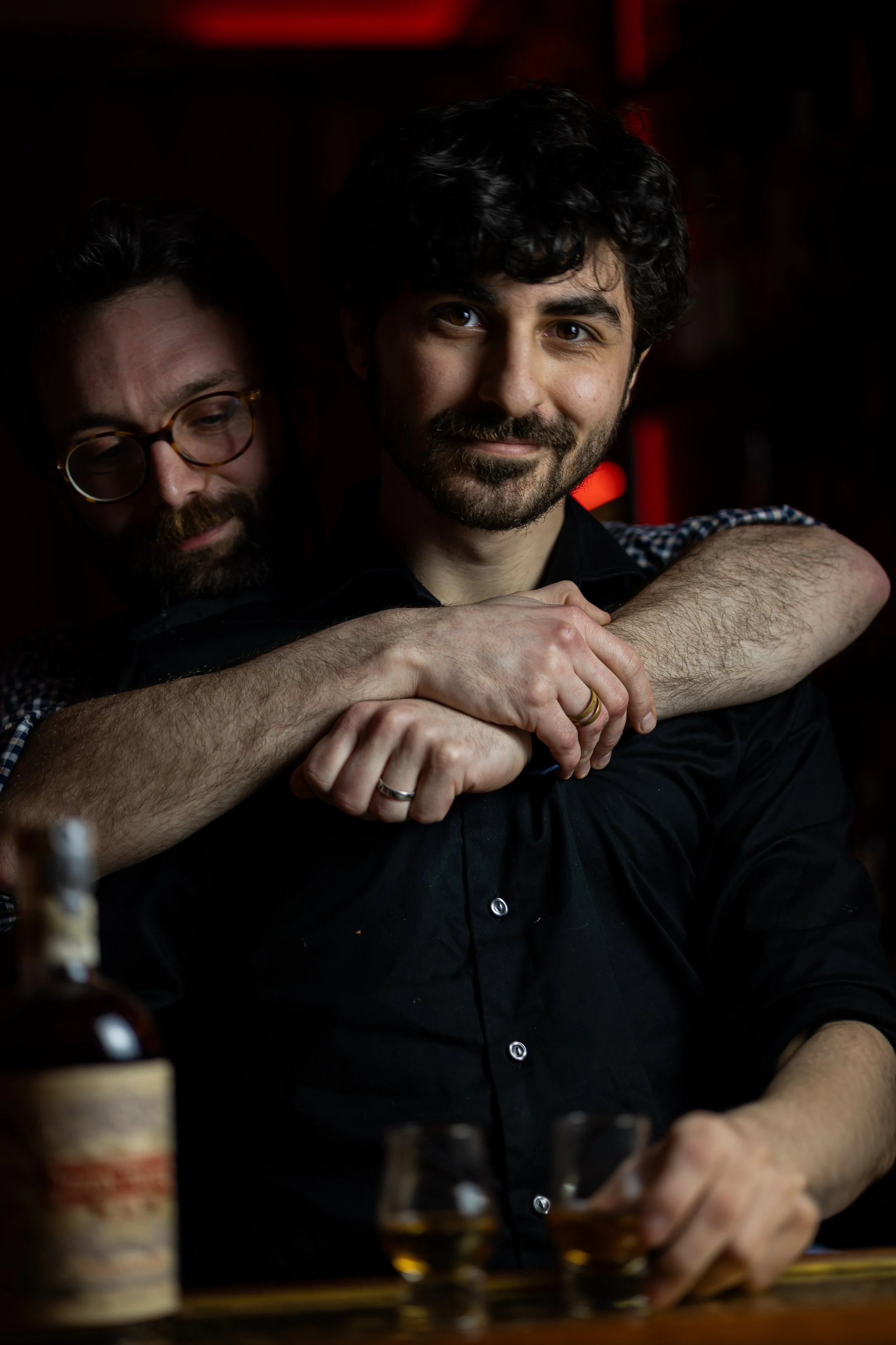 Two men at a bar, one sitting and the other hugging him from behind. The man in front has dark curly hair, a beard, and is smiling at the camera. The man behind has glasses, a beard, and is making a playful expression. There are bottles and glasses o