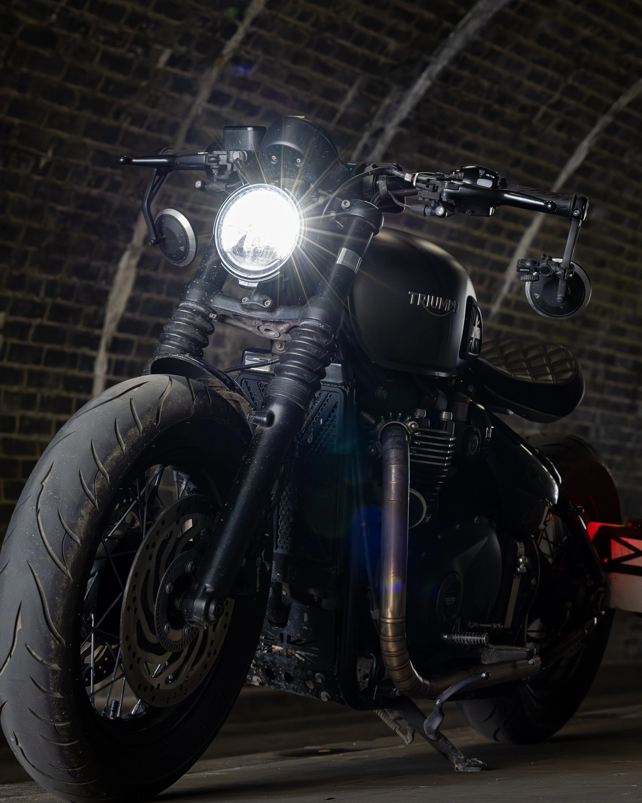 Black Triumph motorcycle with headlight on, parked in a brick tunnel.
