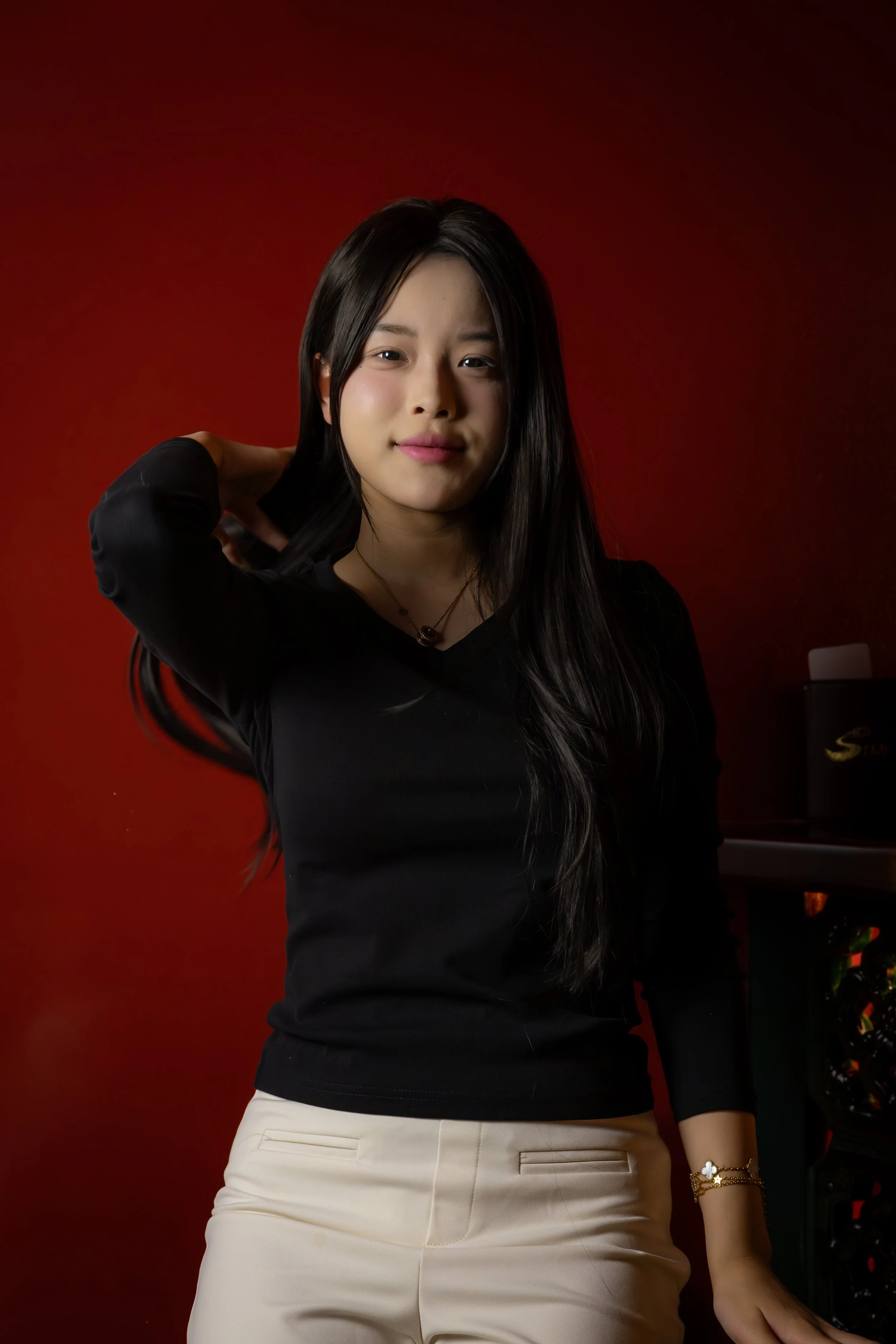 A young woman with long black hair wearing a black long-sleeve shirt and beige pants, standing against a red background, smiling softly with one hand behind her head.