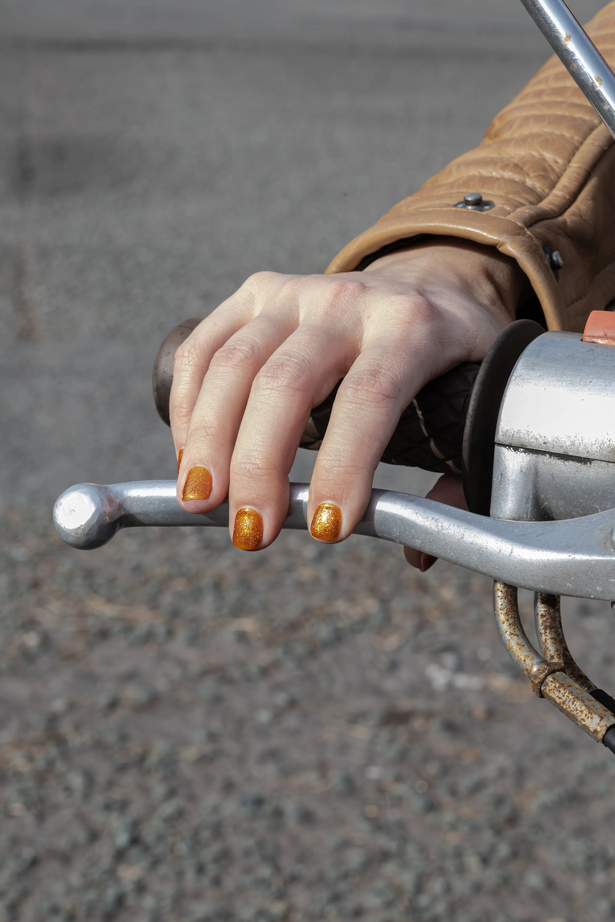 Close-up of a person's hand with orange glitter nail polish gripping a motorcycle handlebar, wearing a brown leather jacket.