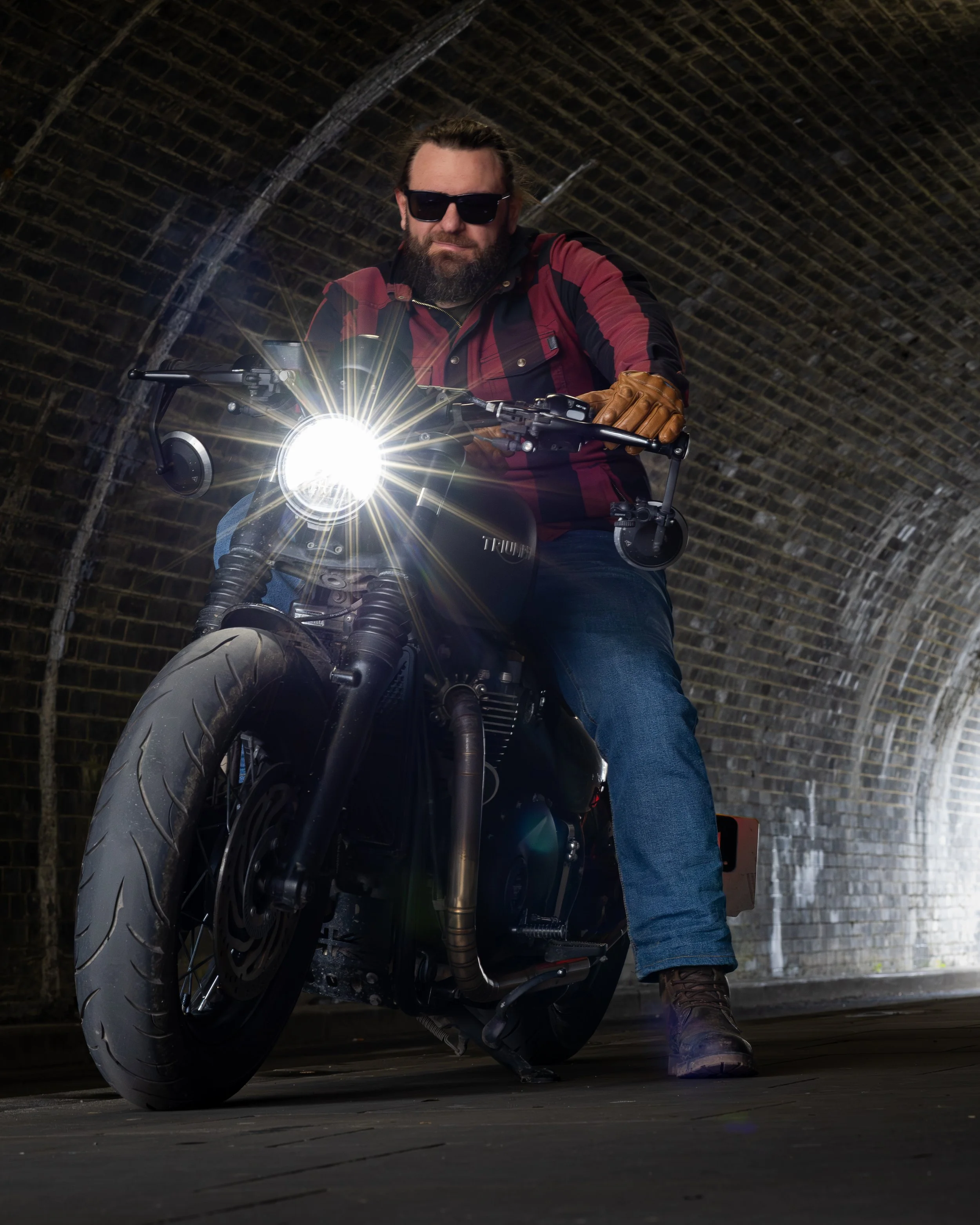 A man riding a black motorcycle through a tunnel with a brick ceiling. He is wearing a red and black jacket, jeans, sunglasses, and brown gloves. The motorcycle's headlight is on, creating a bright starburst effect.