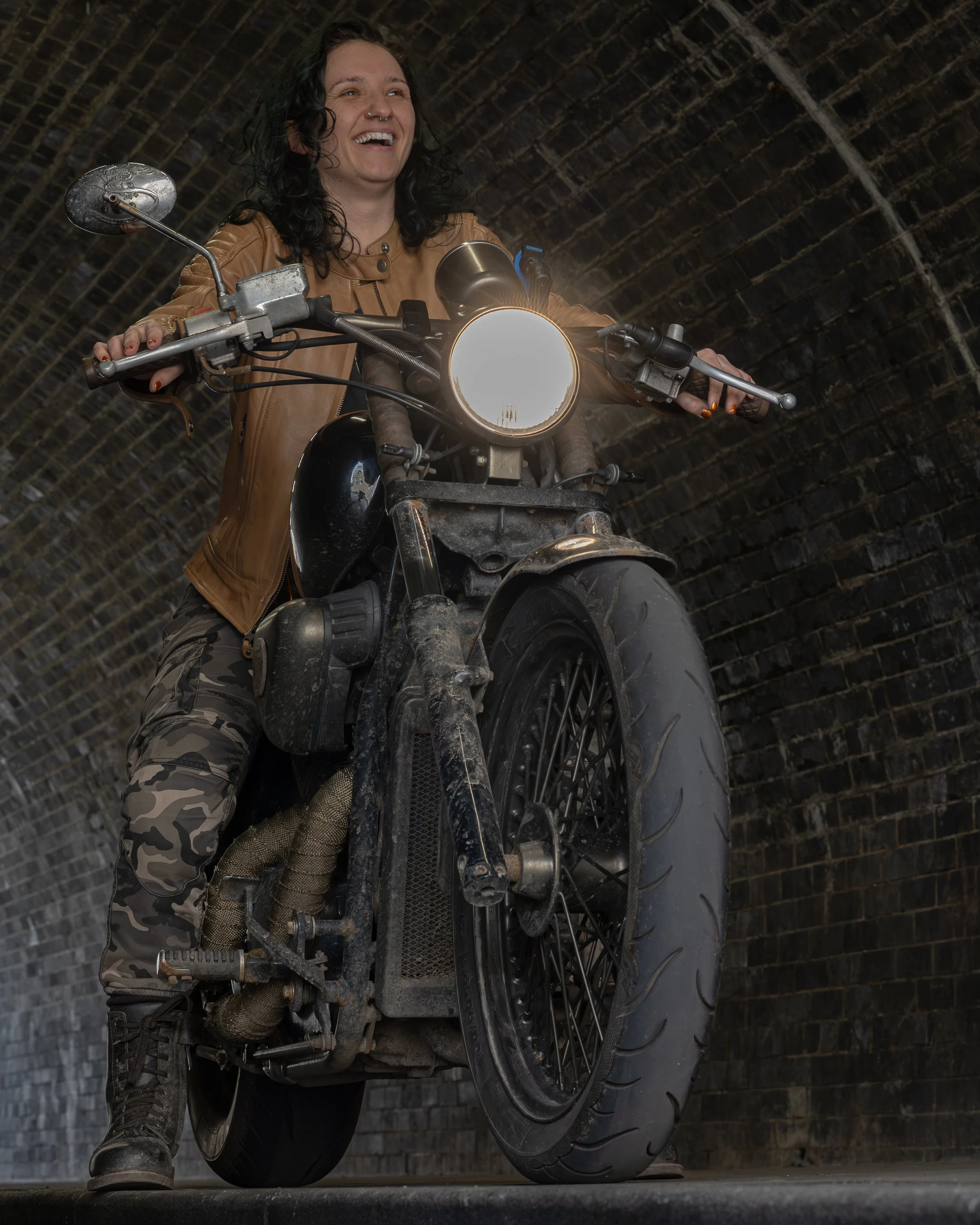 A woman riding a black motorcycle through a dark tunnel, smiling, wearing a tan jacket and camouflage pants.