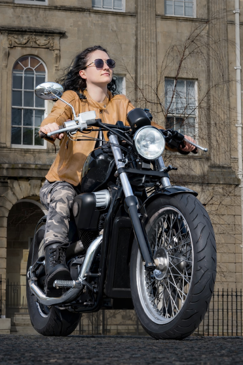 Woman with curly dark hair wearing sunglasses and a brown leather jacket riding a black motorcycle in front of a stone building with arched windows and decorative elements.
