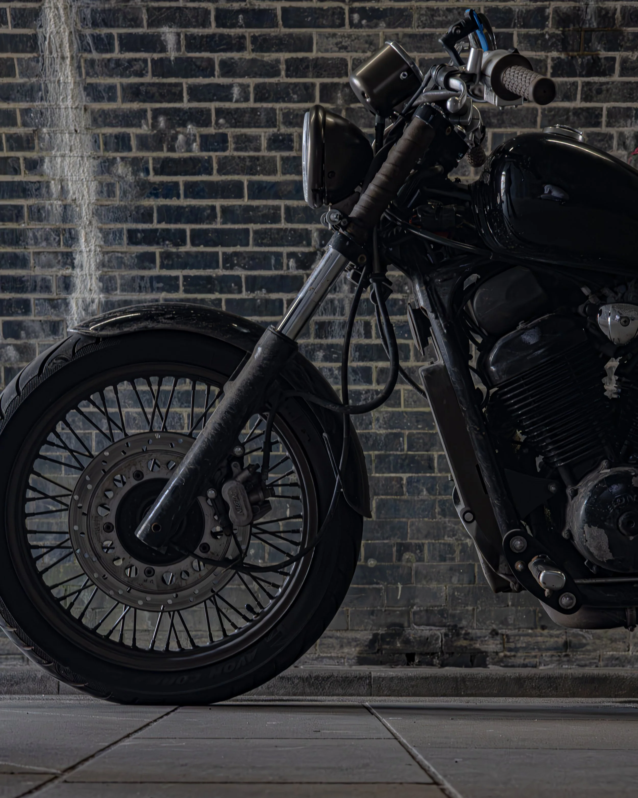 Close-up of a black motorcycle against a brick wall, showing the front wheel, brake disc, suspension, and part of the engine.