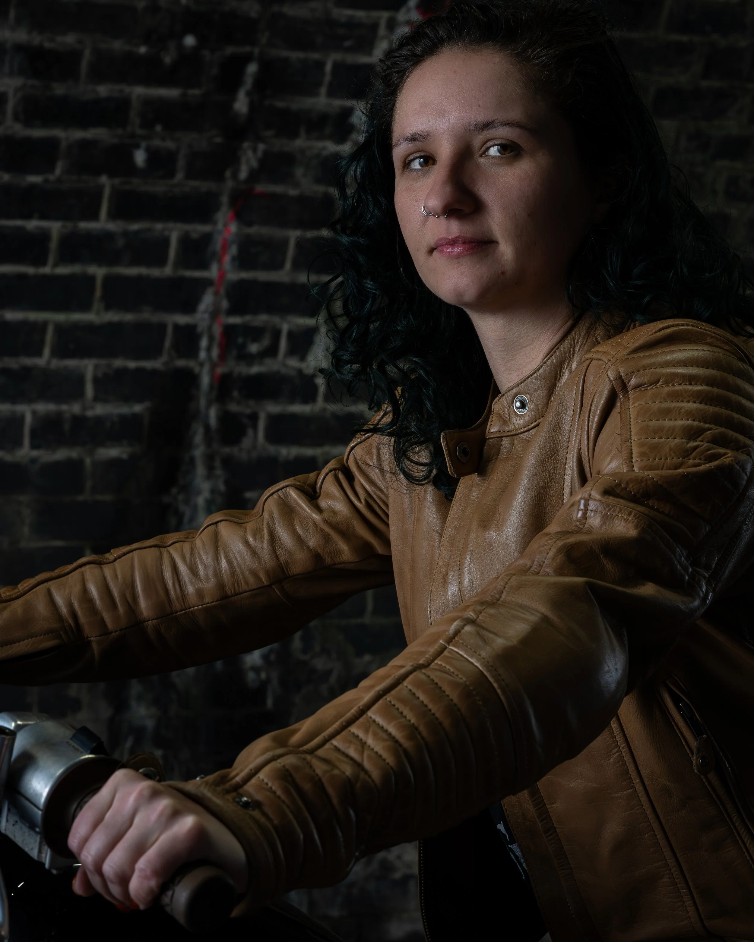 A young woman with black, curly hair and a septum piercing wearing a brown leather jacket riding a motorcycle against a dark brick wall background.