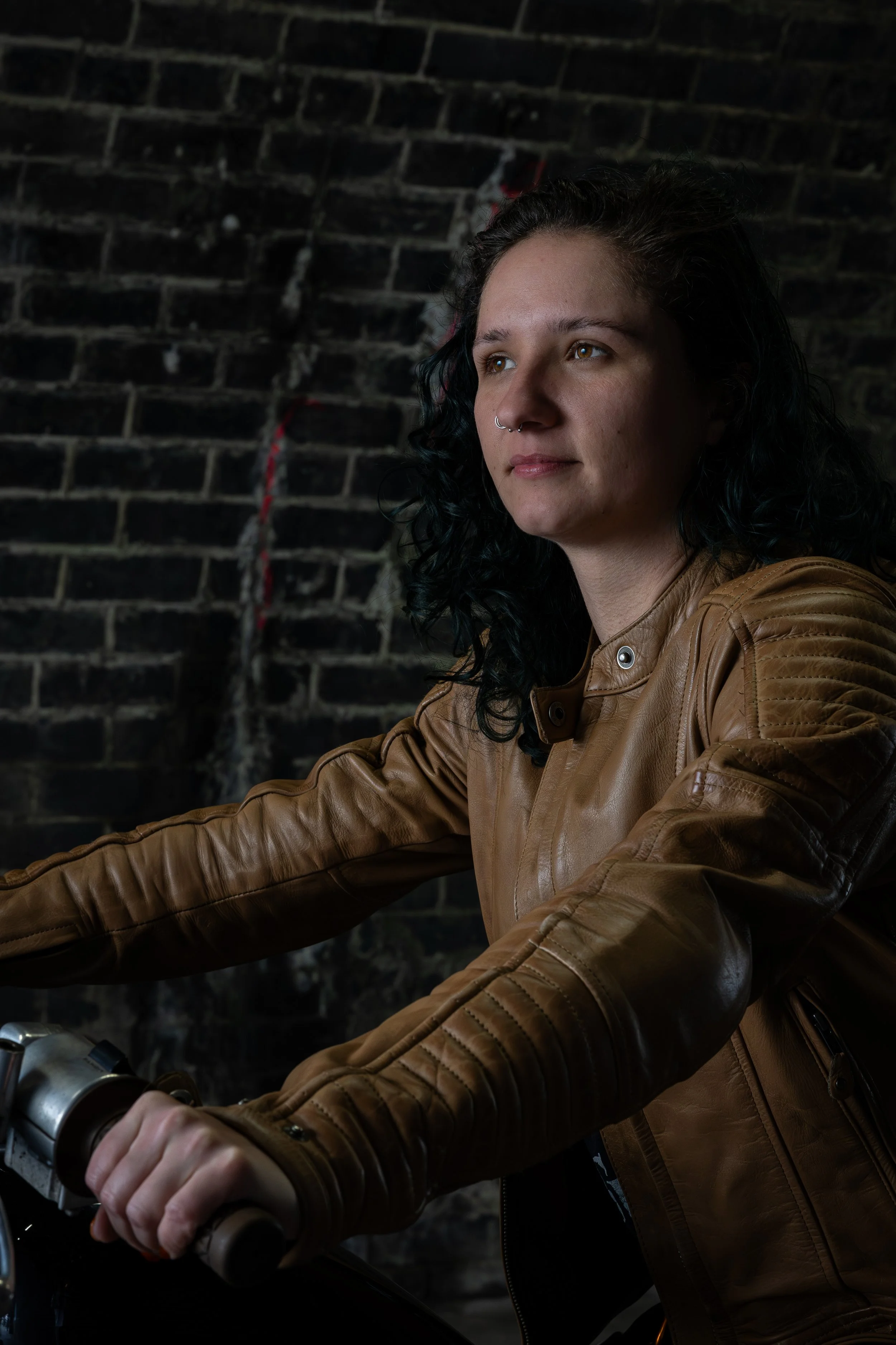 A young woman with curly dark hair, wearing a brown leather jacket, sitting on a motorcycle against a dark brick wall.