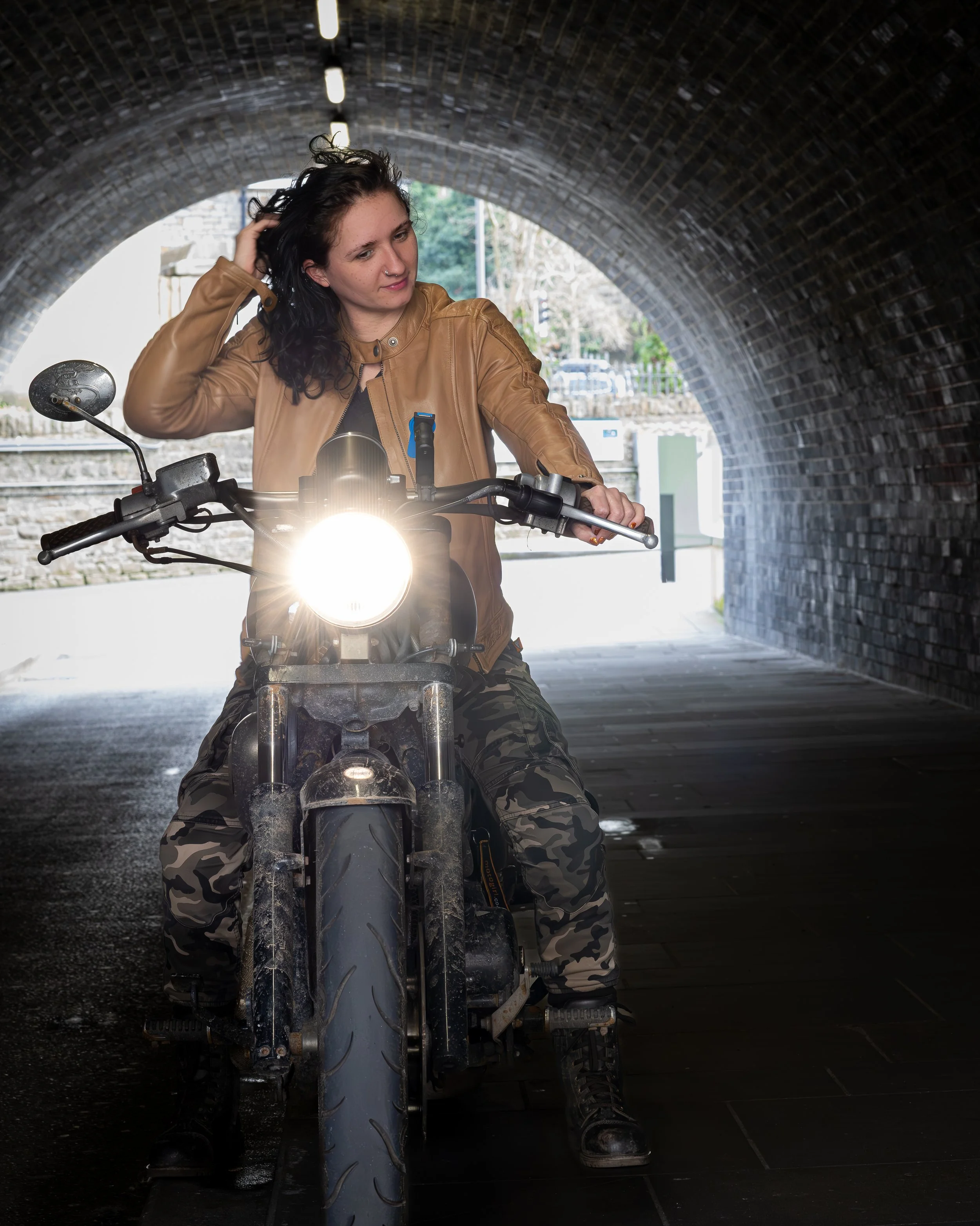 A woman in a brown leather jacket and camouflage pants sits on a black motorcycle inside a dark tunnel, with the motorcycle's headlight turned on.