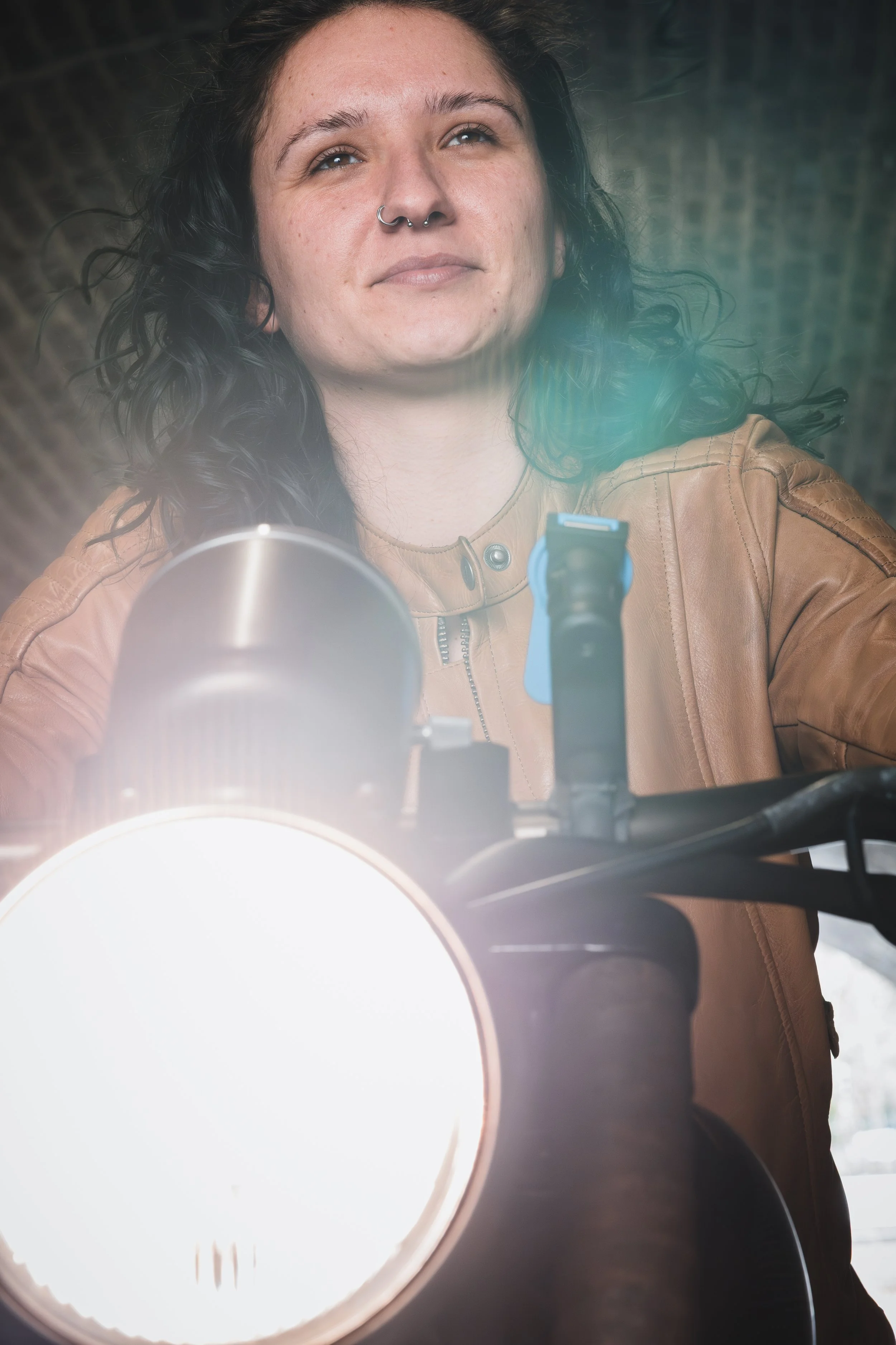 A woman with curly black hair, wearing a tan leather jacket, is taking a selfie with a motorcycle.