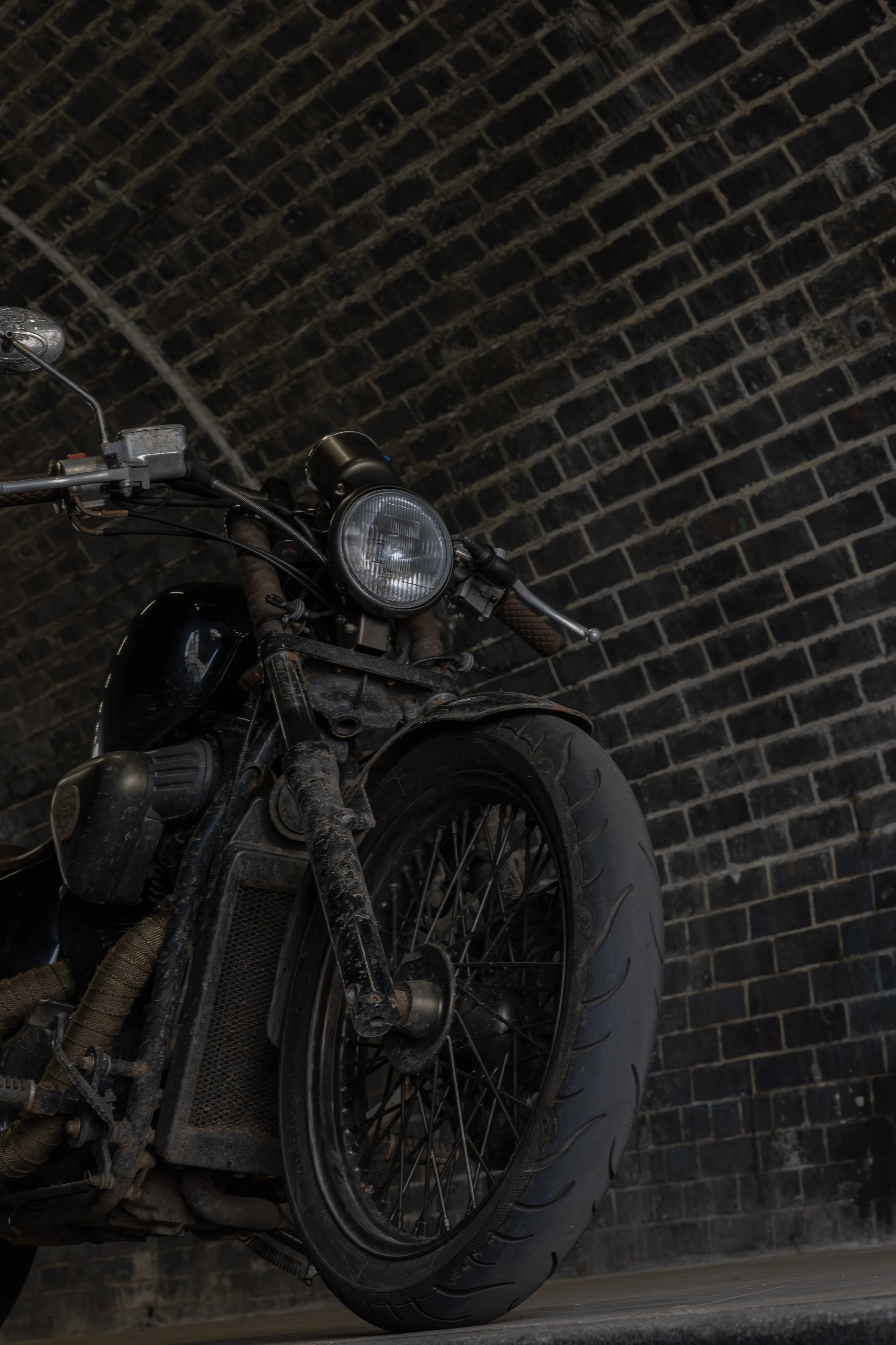 An old, dusty black motorcycle with a large front tire, parked against a dark brick wall.