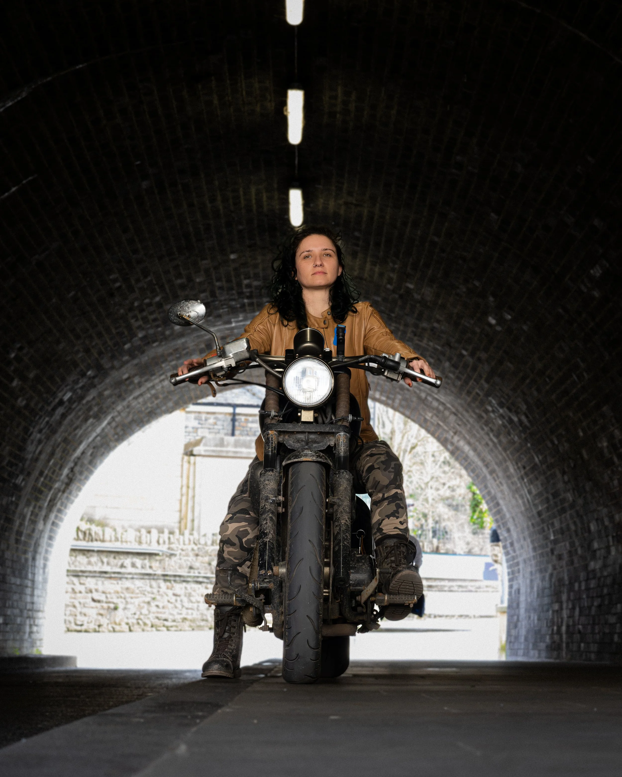 A woman riding a motorcycle through a tunnel, facing forward with serious expression, wearing a brown leather jacket and camouflage pants.