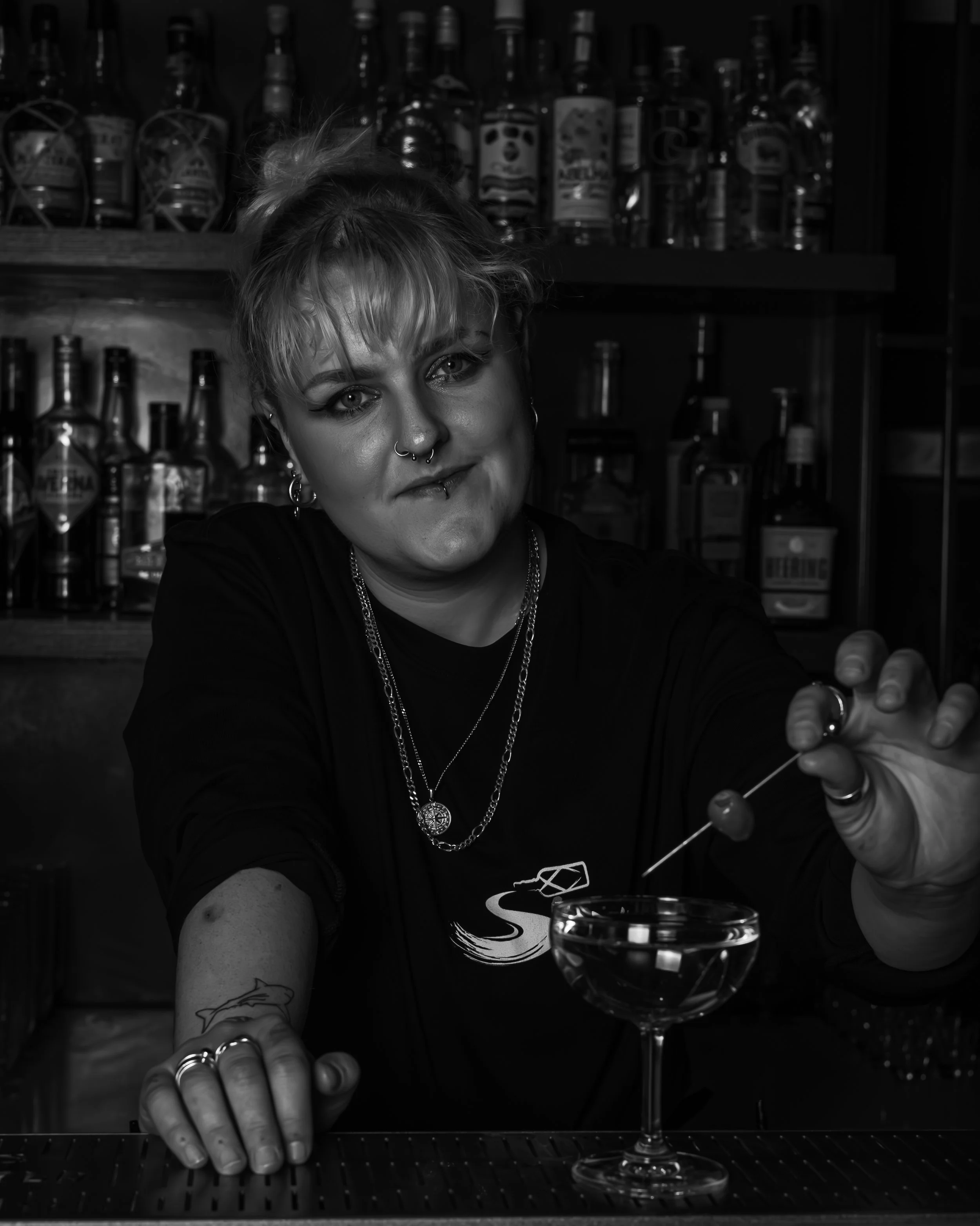 A woman with short blonde hair and multiple jewelry pieces, including rings and necklaces, is preparing a cocktail with an eyedropper at a bar. She has a septum piercing and tattoos, and is smiling slightly. Behind her are shelves with bottles of alc