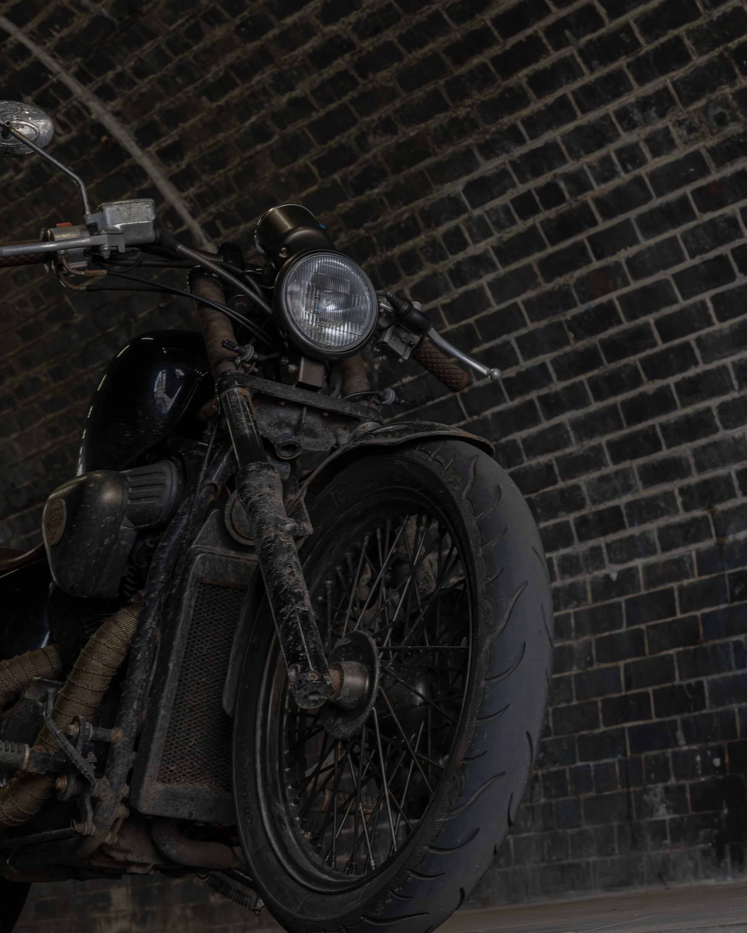 A vintage black motorcycle with a large front tire, round headlight, and rusty parts, leaning against a dark brick wall.