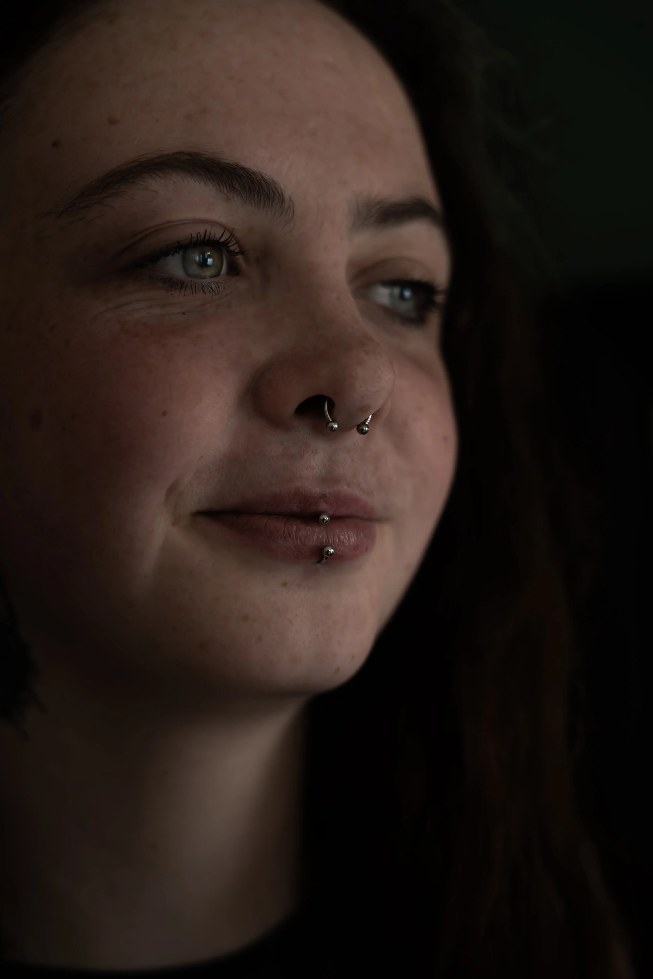 Un portrait d'une femme avec des piercings au nez et à la lèvre, avec les yeux bleus, sur fond sombre.