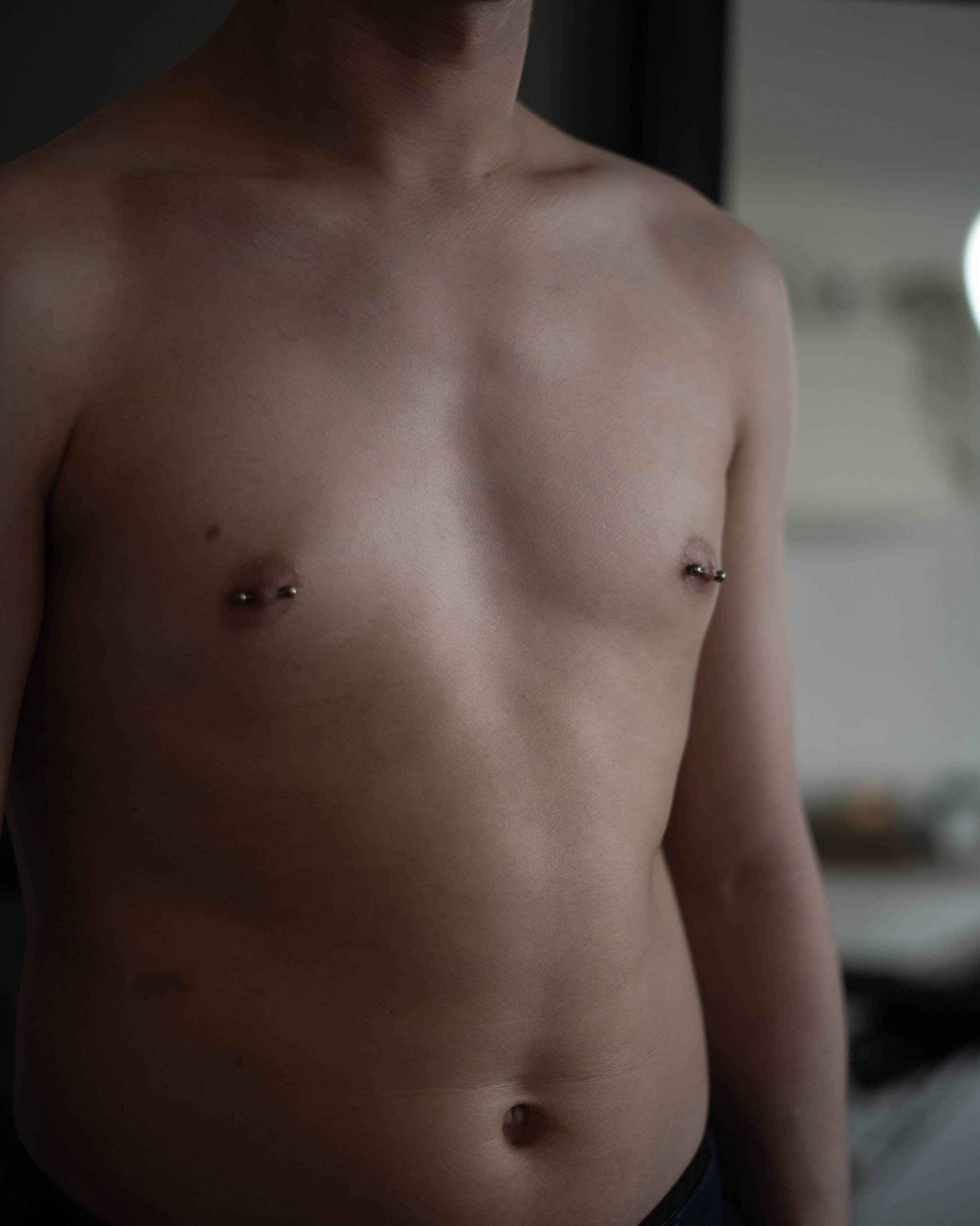 Un homme torse nu avec des piercings au niveau des mamelons, pris de face dans une pièce intérieure.