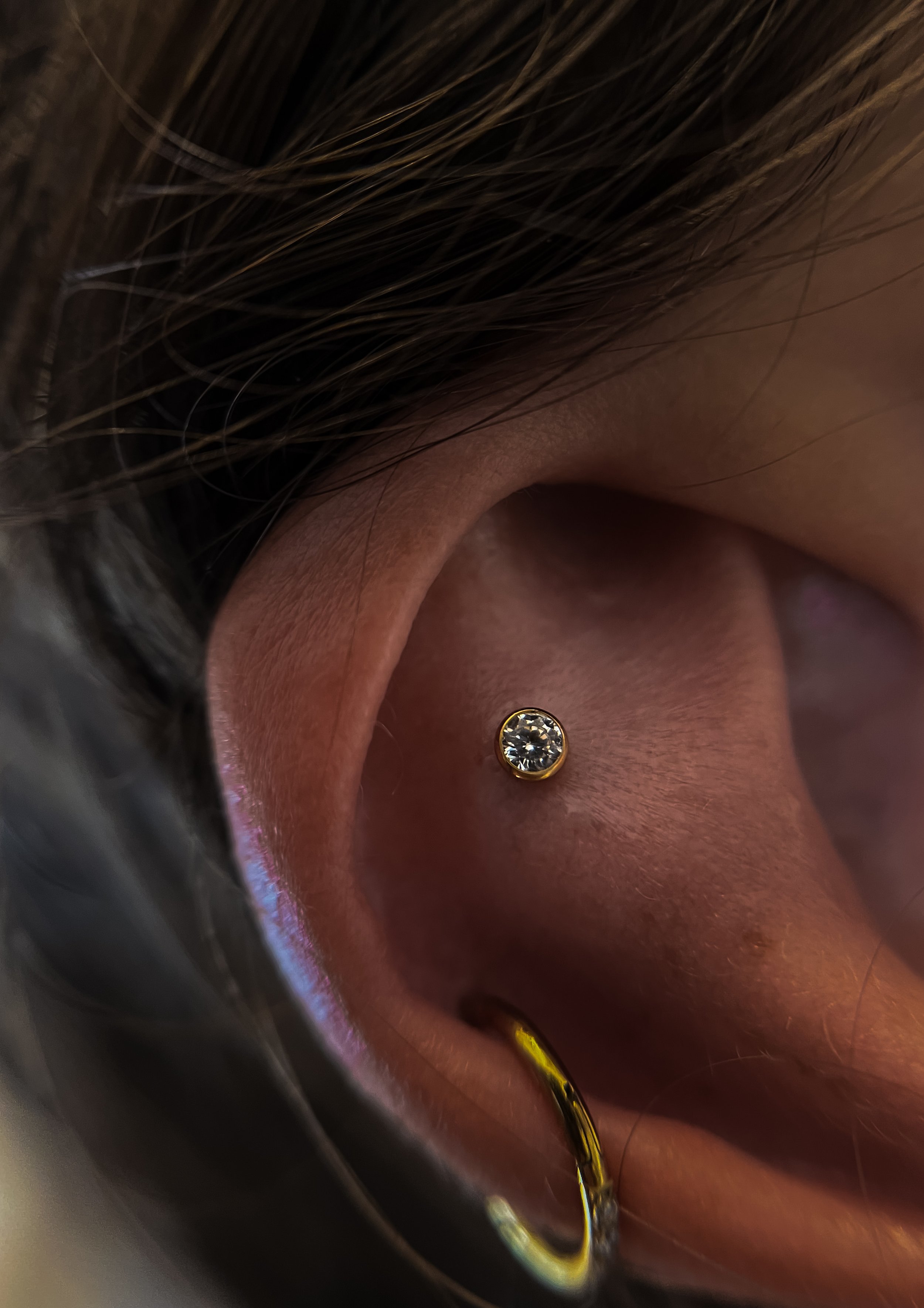 Zoom sur un piercing à la tragus avec une pierre précieuse entourée d'un cadre en or dans l'oreille d'une personne.