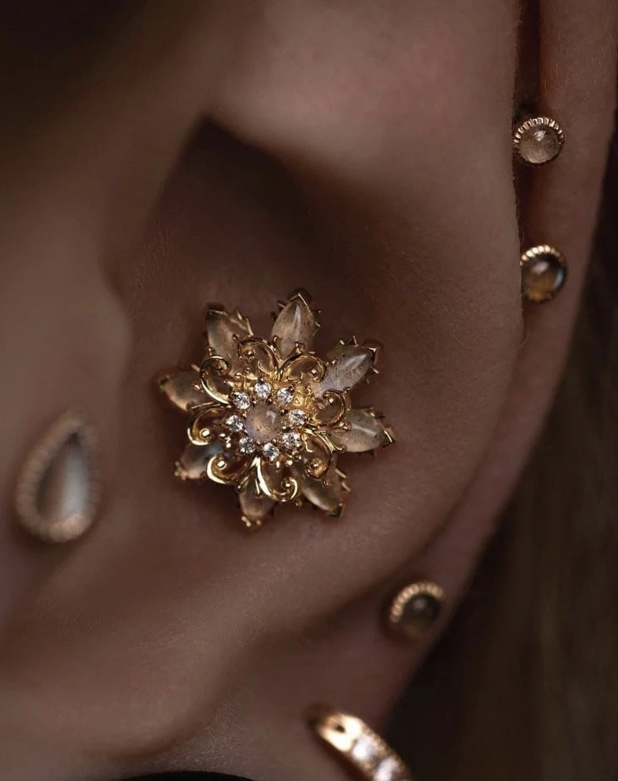 Close-up d'une oreille ornée de multiples bijoux en or, y compris une broche florale avec des pierres précieuses et des diamants, ainsi que des petites boucles d'oreilles et un anneau.