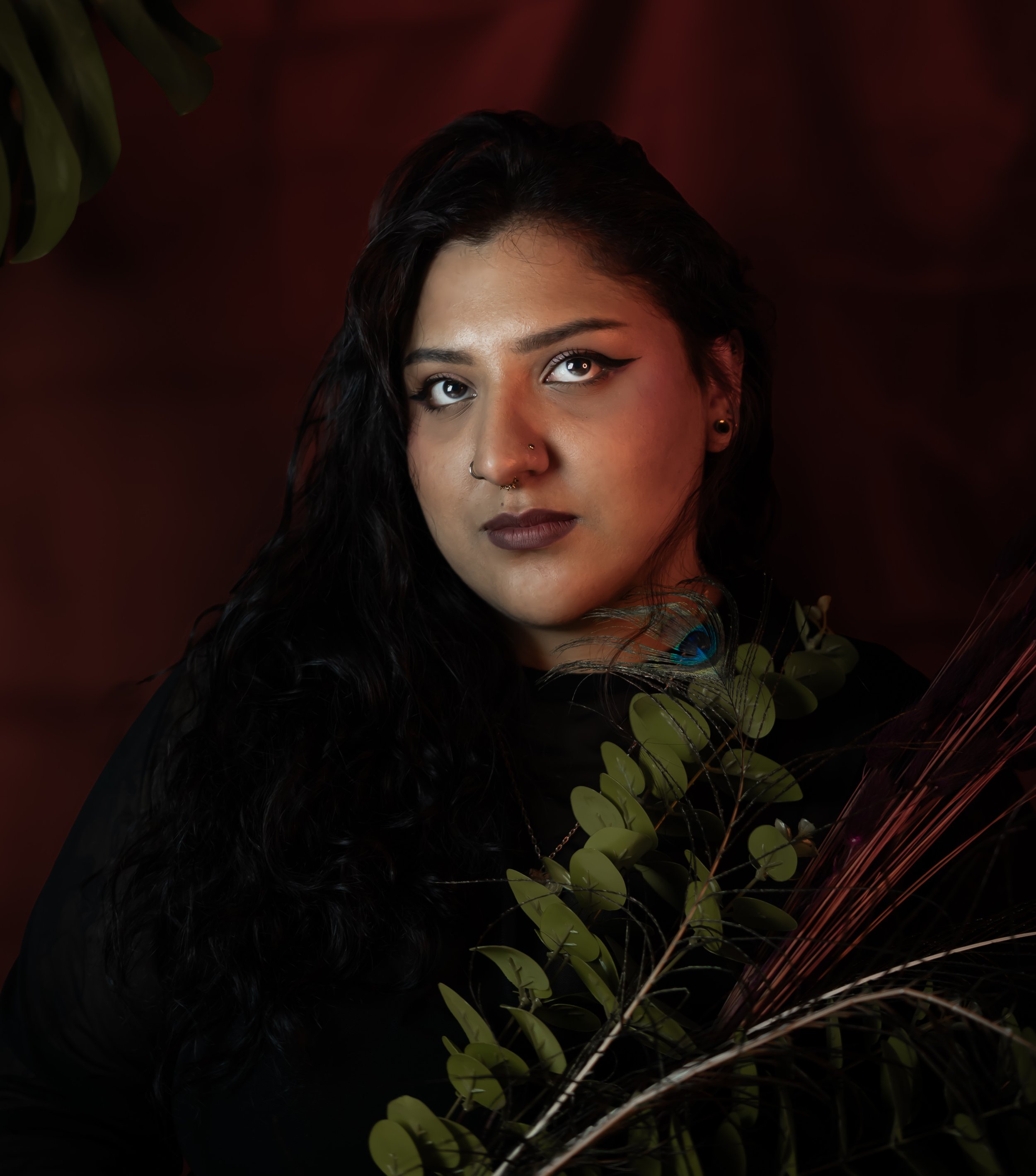 Portrait d'une femme aux cheveux noirs, portant du maquillage foncé et des piercings, entourée de plantes vertes