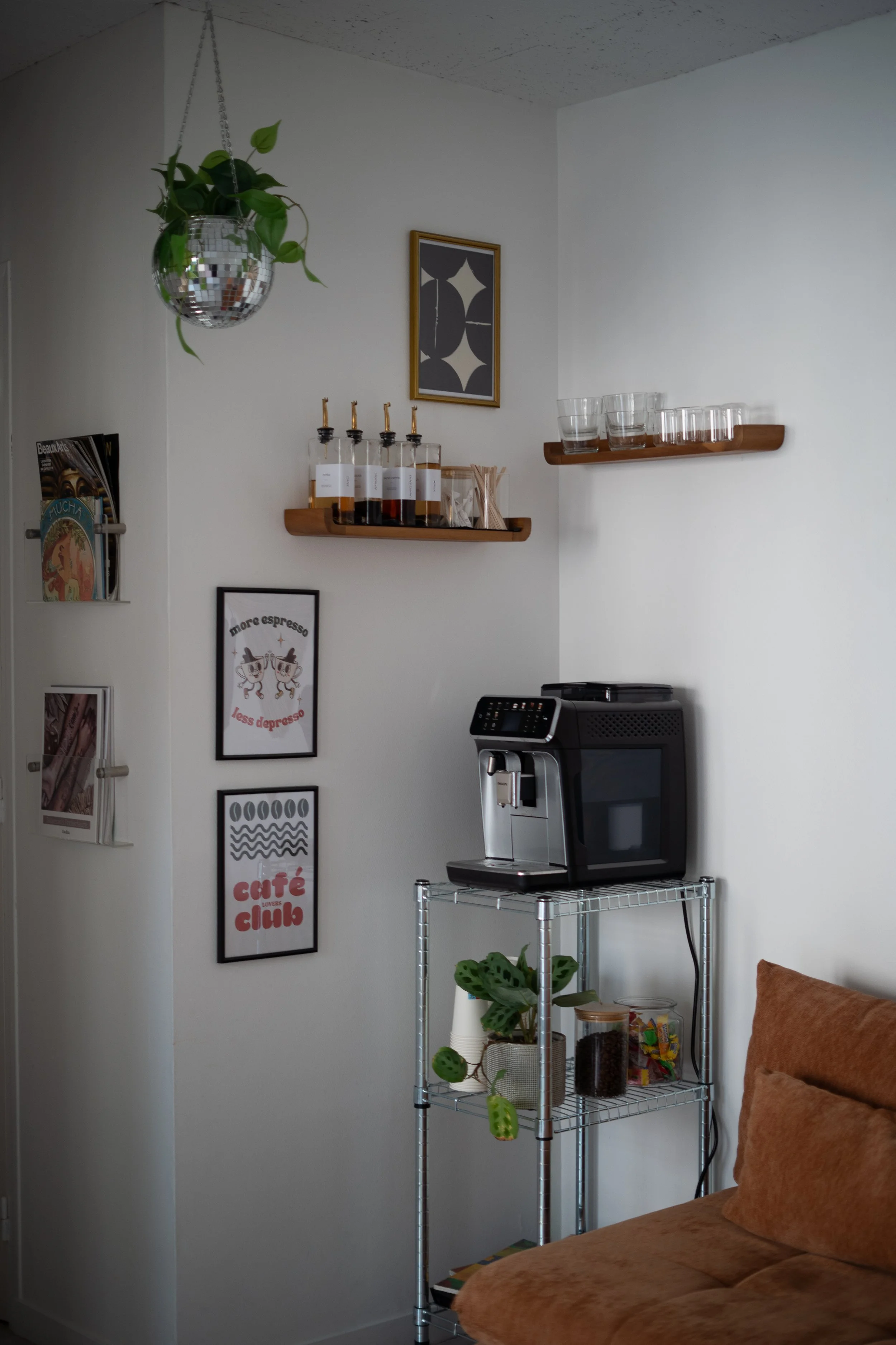 Coin de salon avec une étagère en métal contenant une machine à café, une plante, des bocaux avec des épices ou café, et un canapé marron. Deux autres étagères en bois fixées au mur avec des verres et des bouteilles. Mur blanc avec des cadres décoratifs et magazines.