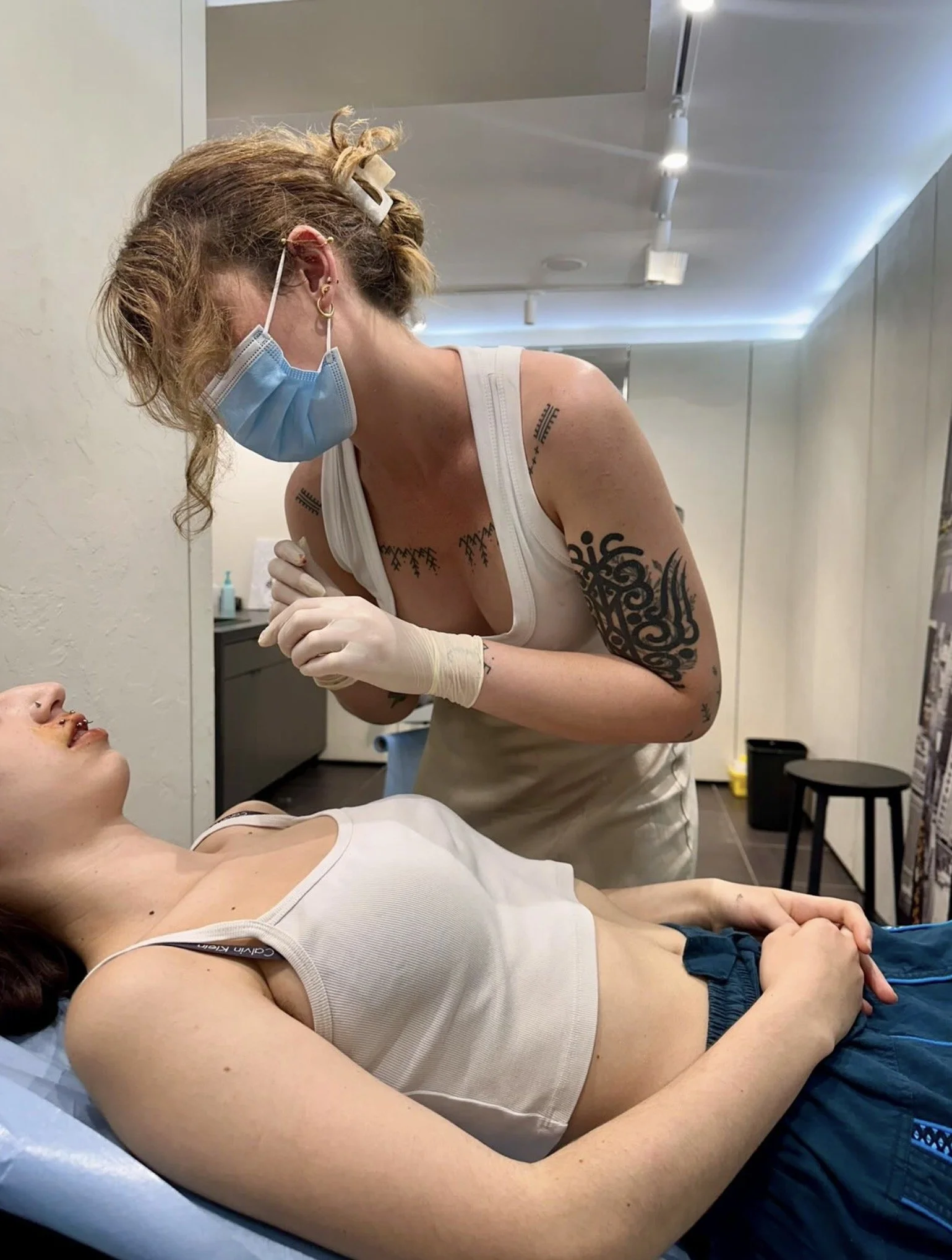 Une femme allongée sur une table lors d'une séance de tatouage, un tatoueur portant un masque, des gants et une robe sans manches travaille sur un tatouage sur le bras de la femme.