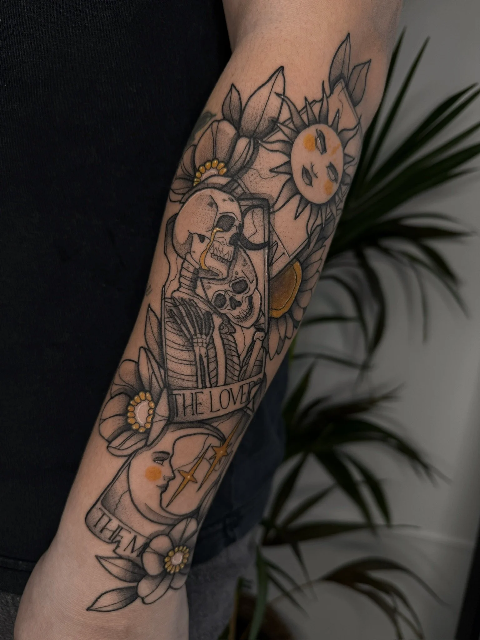 Tatouage représentant une scène avec un soleil avec un visage, des fleurs, deux crânes, une femme et une lune avec un visage, accompagnés de rubans et de la phrase 'THE LOVER'.