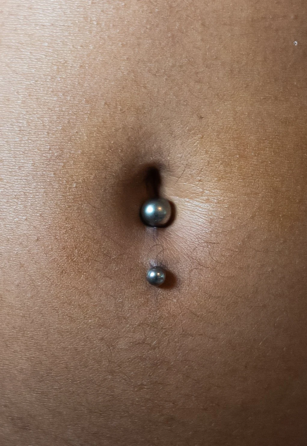 Un piercings inversé au nombril avec deux boules en métal, le ventre d'une personne nue avec une peau de couleur foncée.