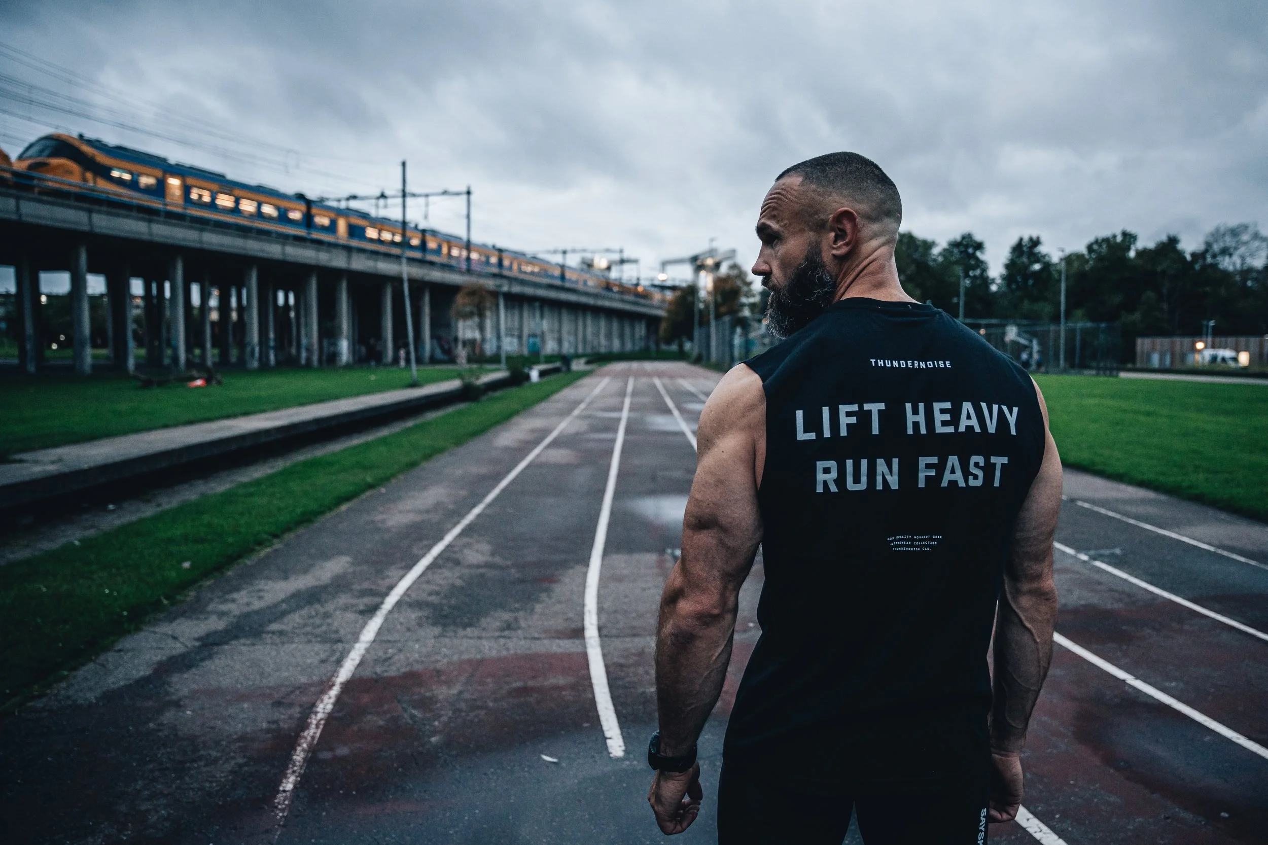A muscular man with a beard, wearing a black sleeveless shirt that says 'Lift Heavy Run Fast,' standing on an outdoor running track under a cloudy sky.