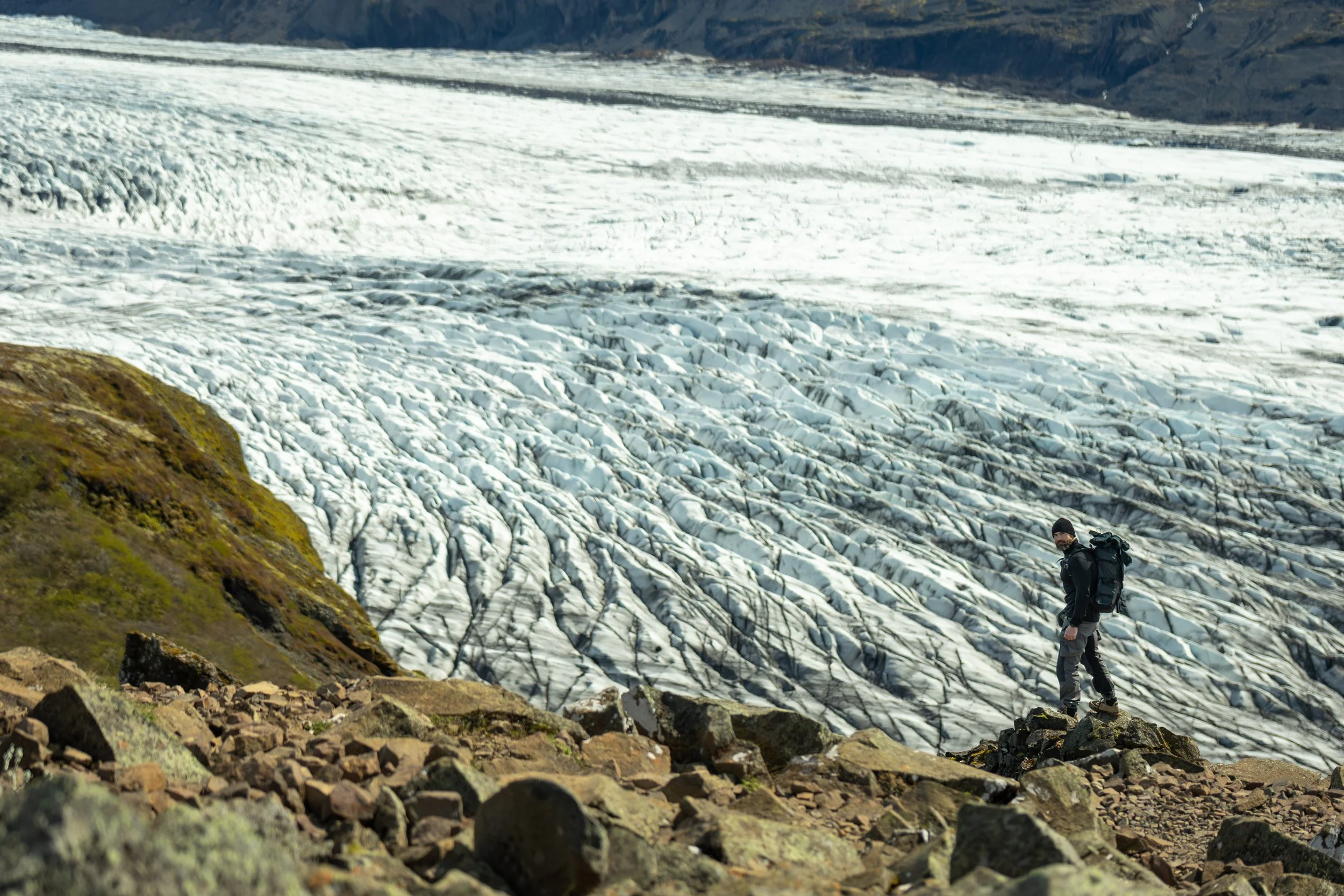 A hiker wearing black clothing and a backpack walking on rocky terrain with a glacier in the background.