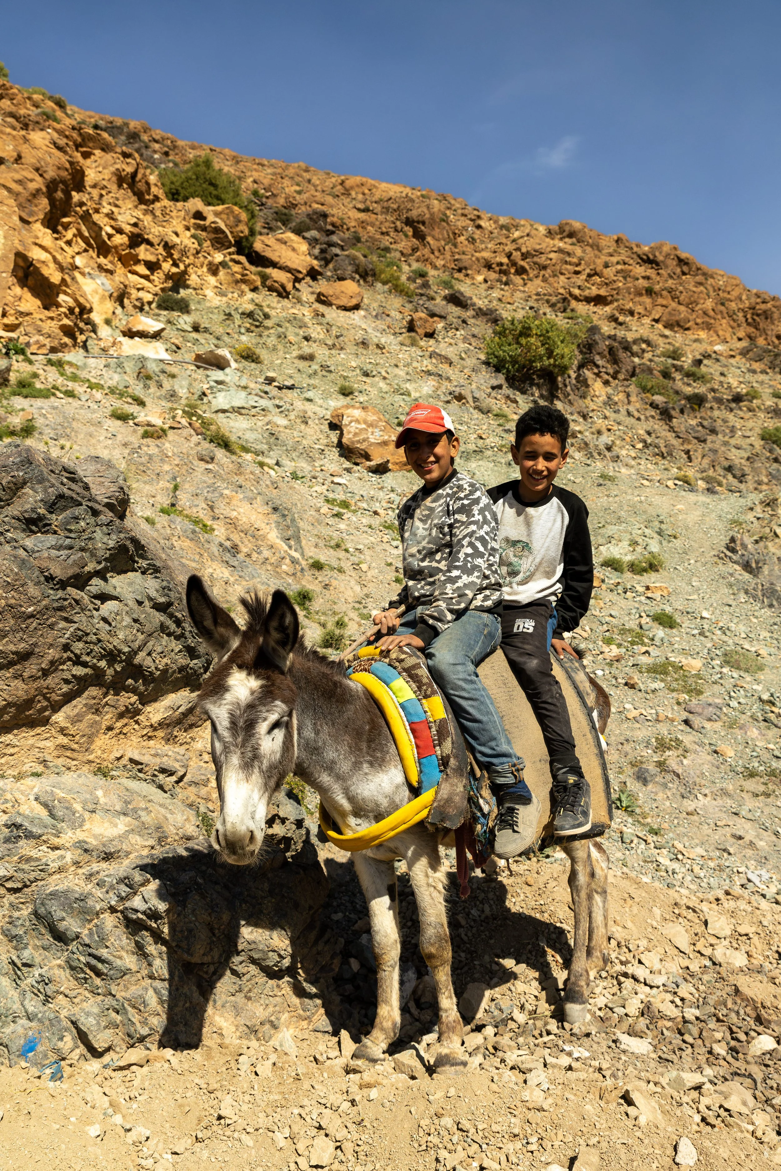 Two young boys sitting on a donkey in a rocky desert landscape with a steep hill and sparse vegetation under a clear blue sky.