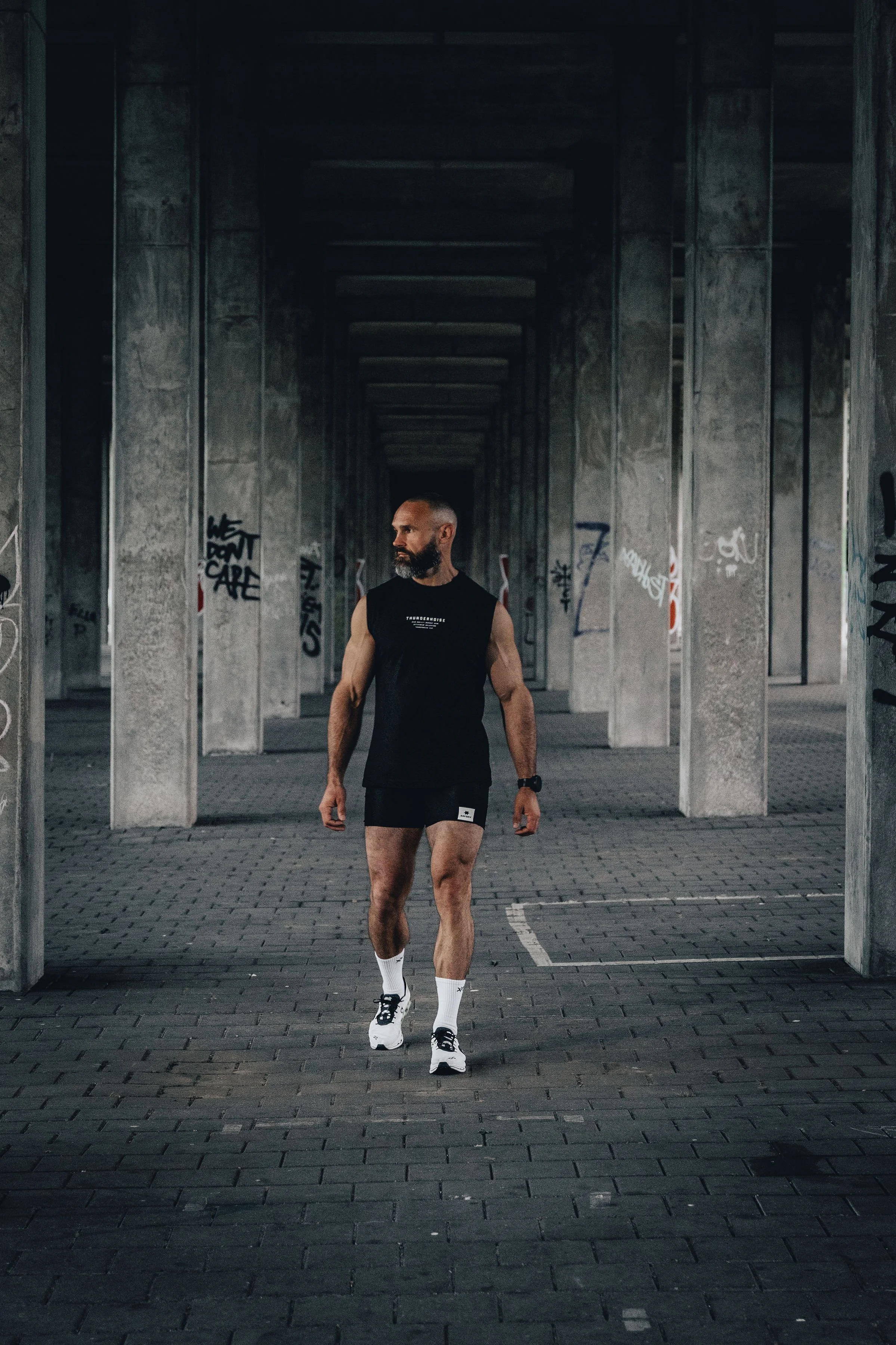 A man wearing a black sleeveless athletic shirt, black shorts, white socks, and white running shoes walking under a concrete bridge with graffiti.