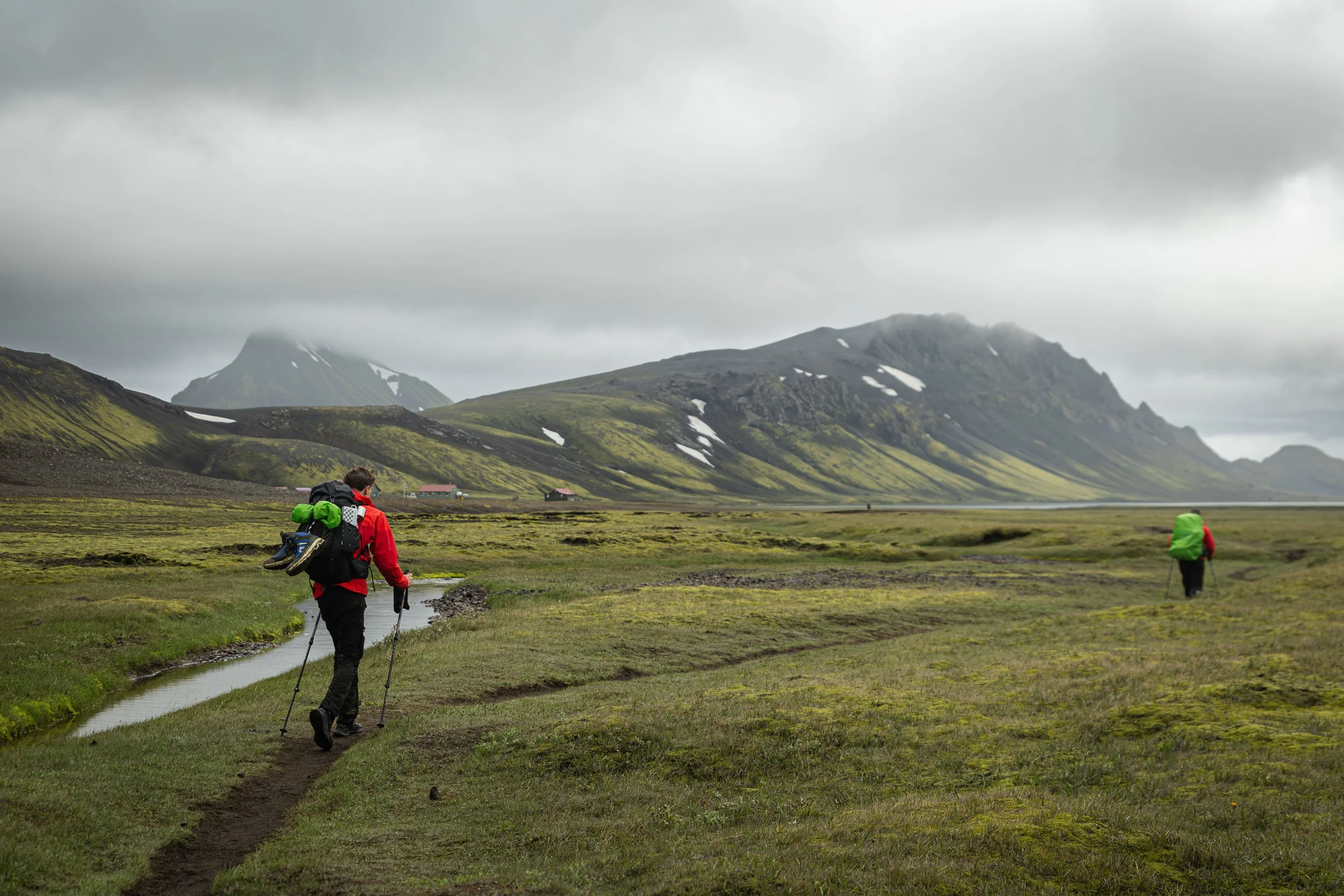 Two hikers dressed in outdoor gear, each with large backpacks, walking along a trail in a lush green landscape with mountains in the background under a cloudy sky.
