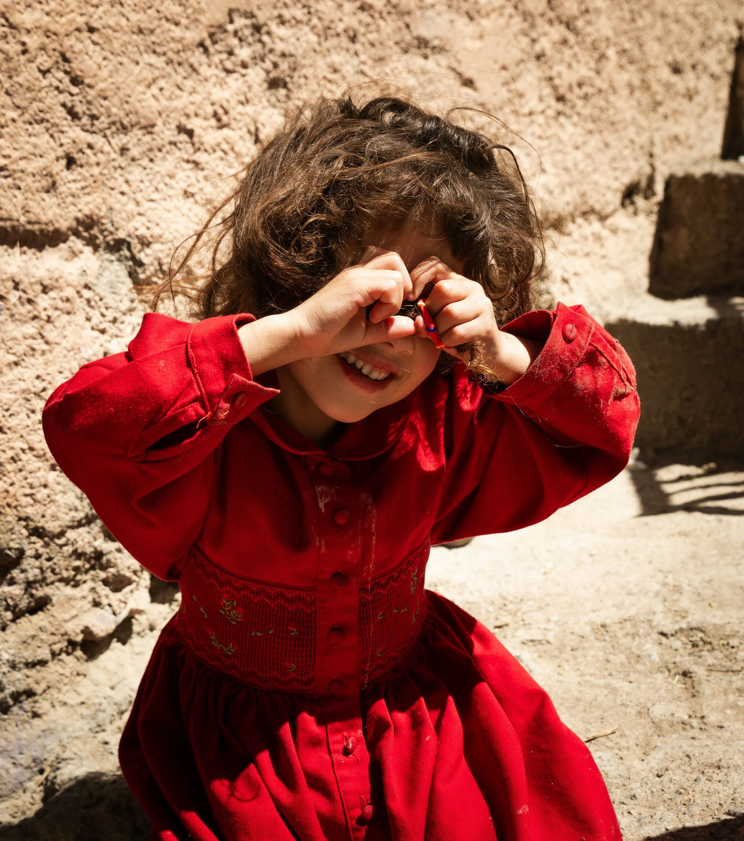 A young girl with curly brown hair wearing a red dress, smiling and making a heart shape with her hands over her eyes, standing outdoors near a stone wall.