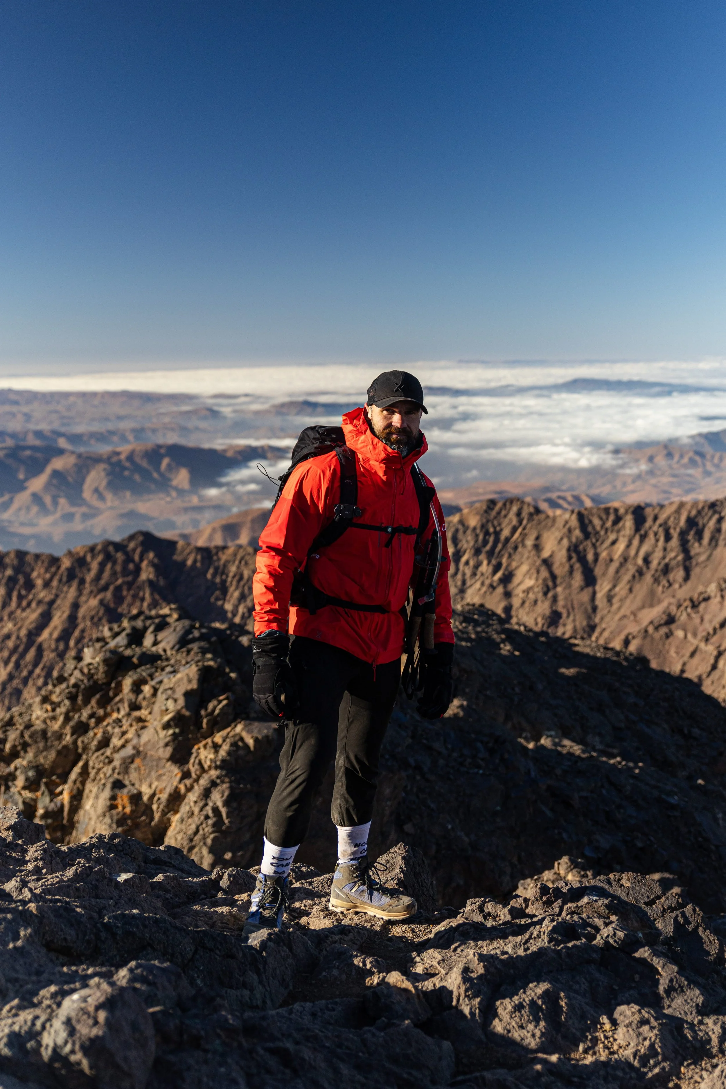 A man in a red jacket, black cap, and hiking gear standing on a rocky mountain summit with a landscape of mountains and clouds in the background.