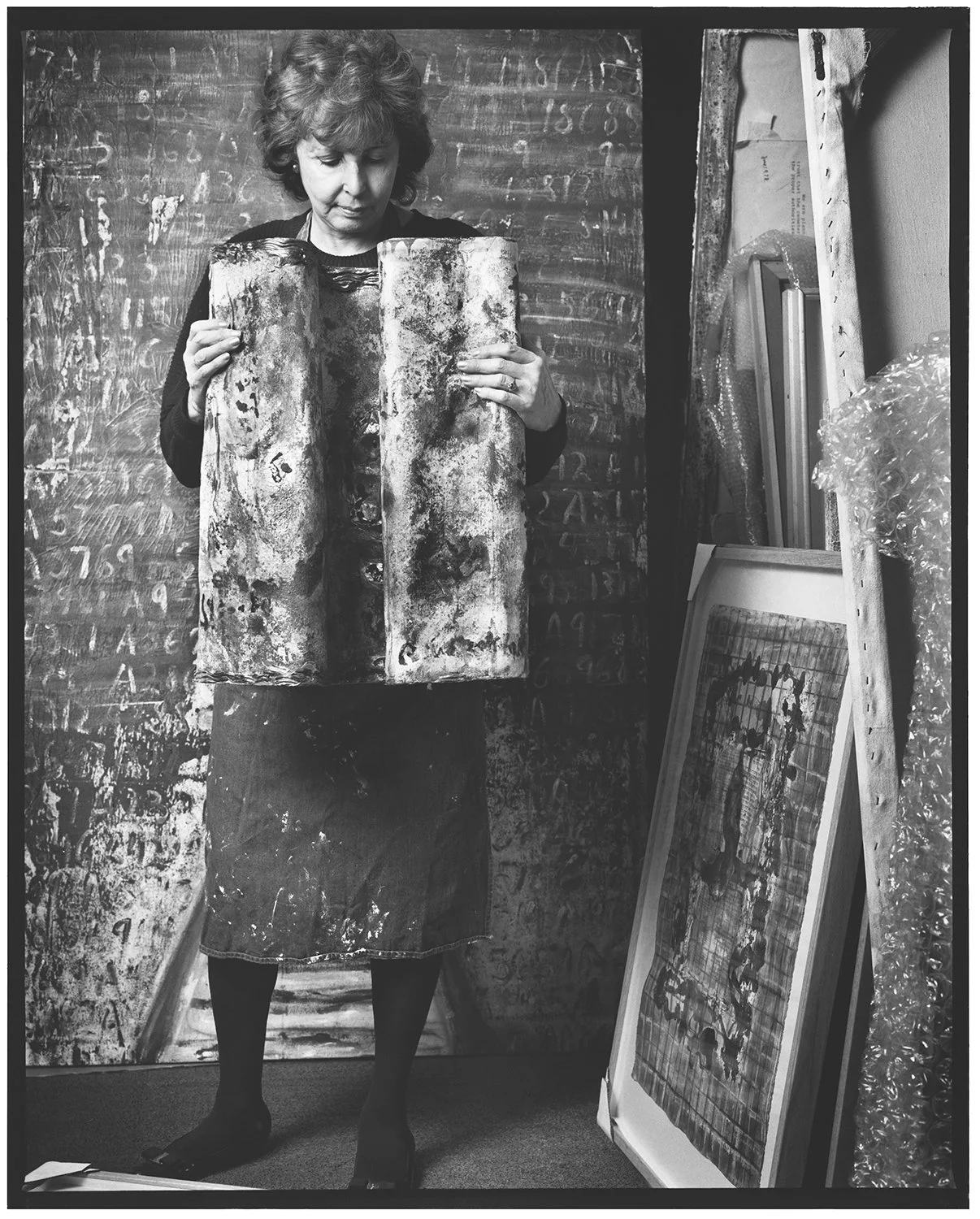 A woman in an art studio holding up two abstract paintings, with additional framed artworks and art supplies on a shelf next to her.