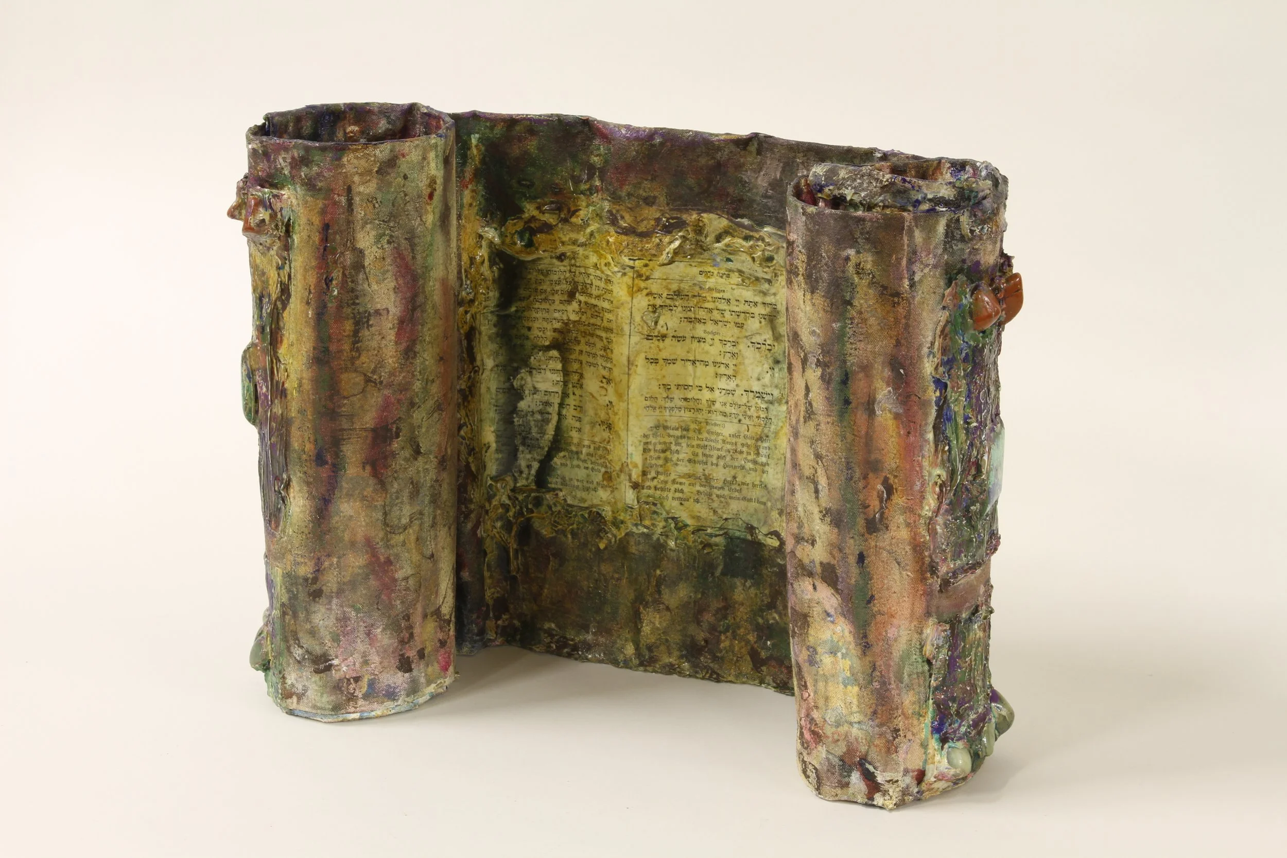 An artistic sculpture resembling an open book with textured, colorful, and irregularly shaped pages, featuring Hebrew text on some pages.