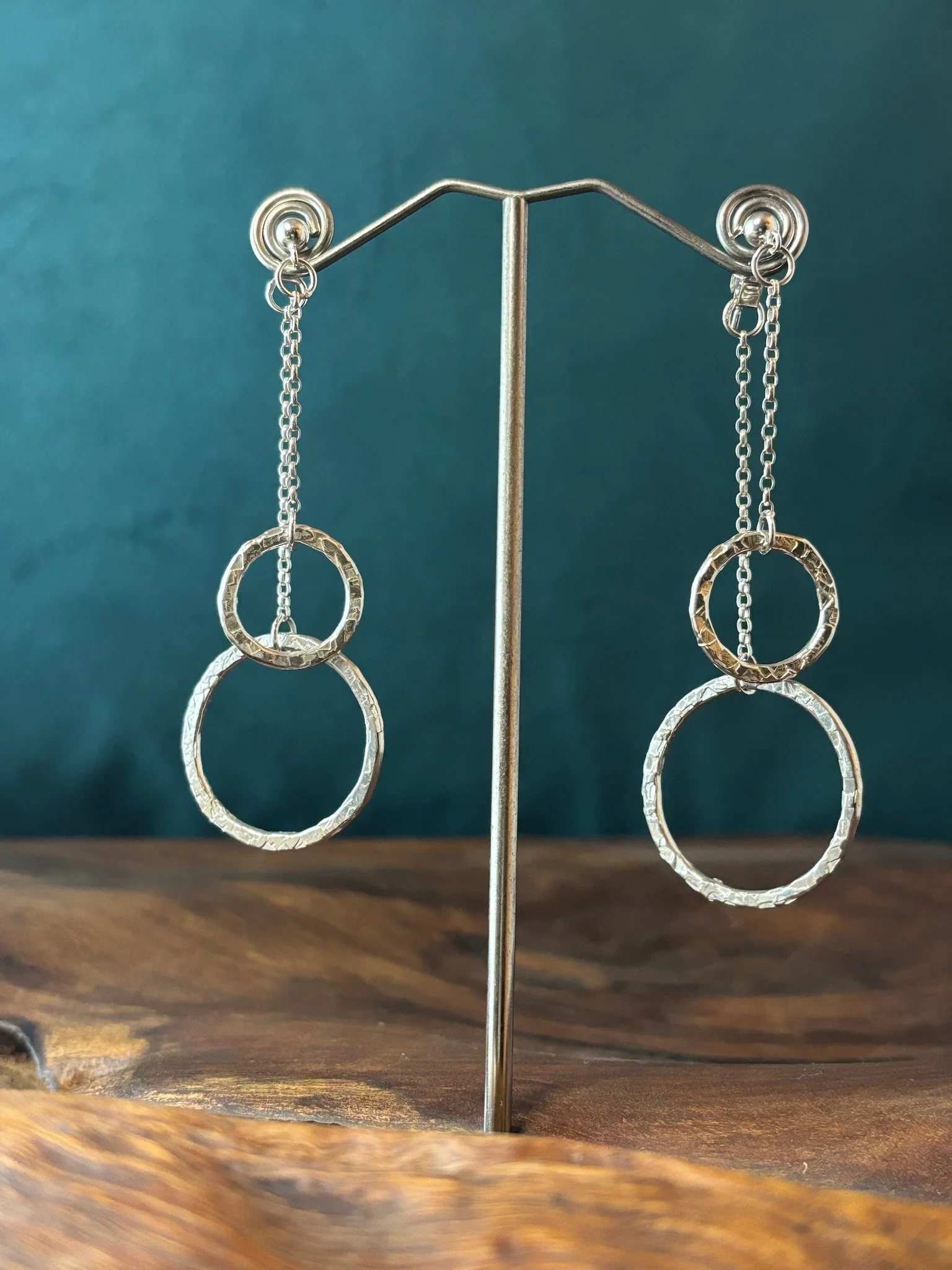 Silver dangling earrings with circular textured links hanging from a jewelry display stand against a wood surface and a teal background.