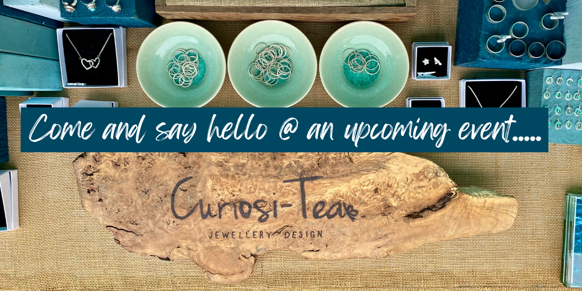 Display of jewelry items including necklaces, rings, and earrings arranged in bowls and boxes on a table at a retail booth, with a sign that reads 'Come and say hello @ an upcoming event' and a wooden sign that says 'Curiosi-Teas Jewelry & Design.'