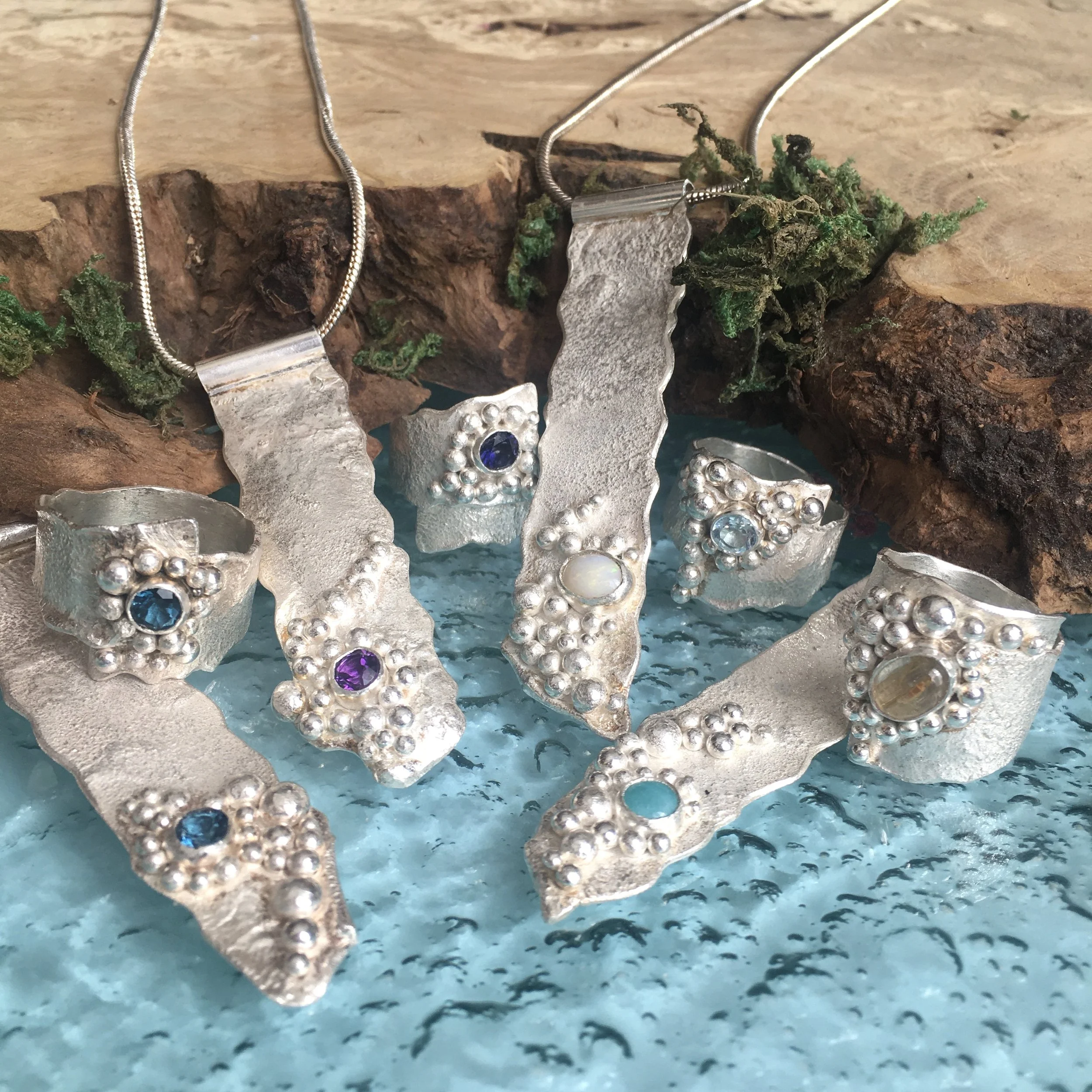 Silver jewelry pieces with blue and white gemstones displayed on a wooden surface with moss and bark.