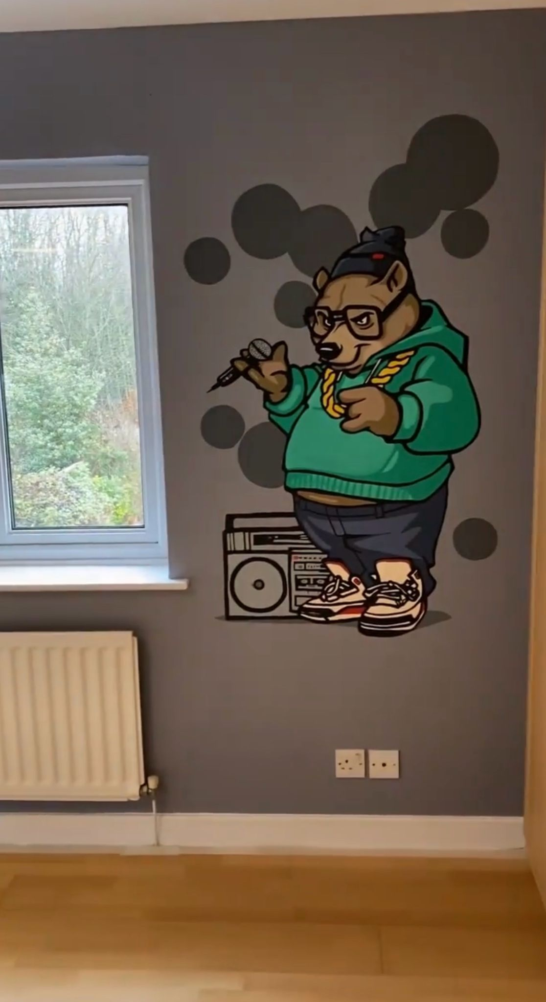 Wall mural of a cartoon bear wearing glasses, a green hoodie, and sneakers, holding a microphone and pointing, with a boombox at its feet and gray circles in the background.