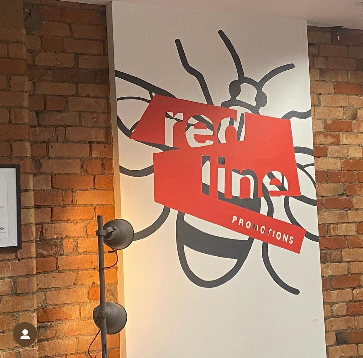 Wall sign displaying the logo and name of 'red line PROMOTIONS' with a beetle illustration in the background, mounted on a brick wall.