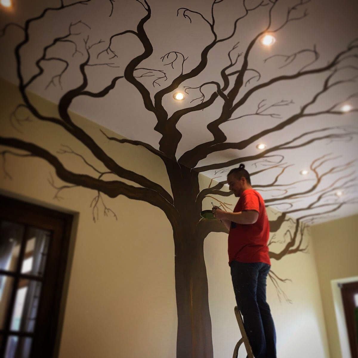 A person standing on a ladder painting a tree silhouette with spreading branches on a ceiling that has built-in lights.