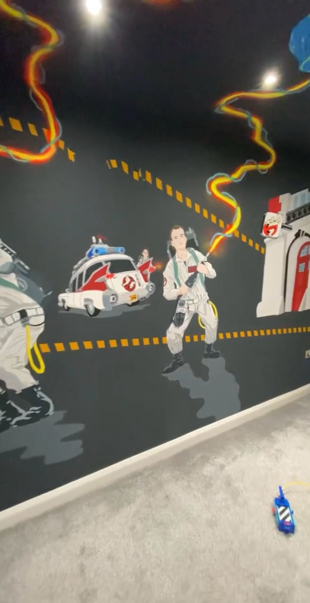 A mural featuring Ghostbusters characters, ghost-hunting equipment, and Ecto-1 car on a dark wall.
