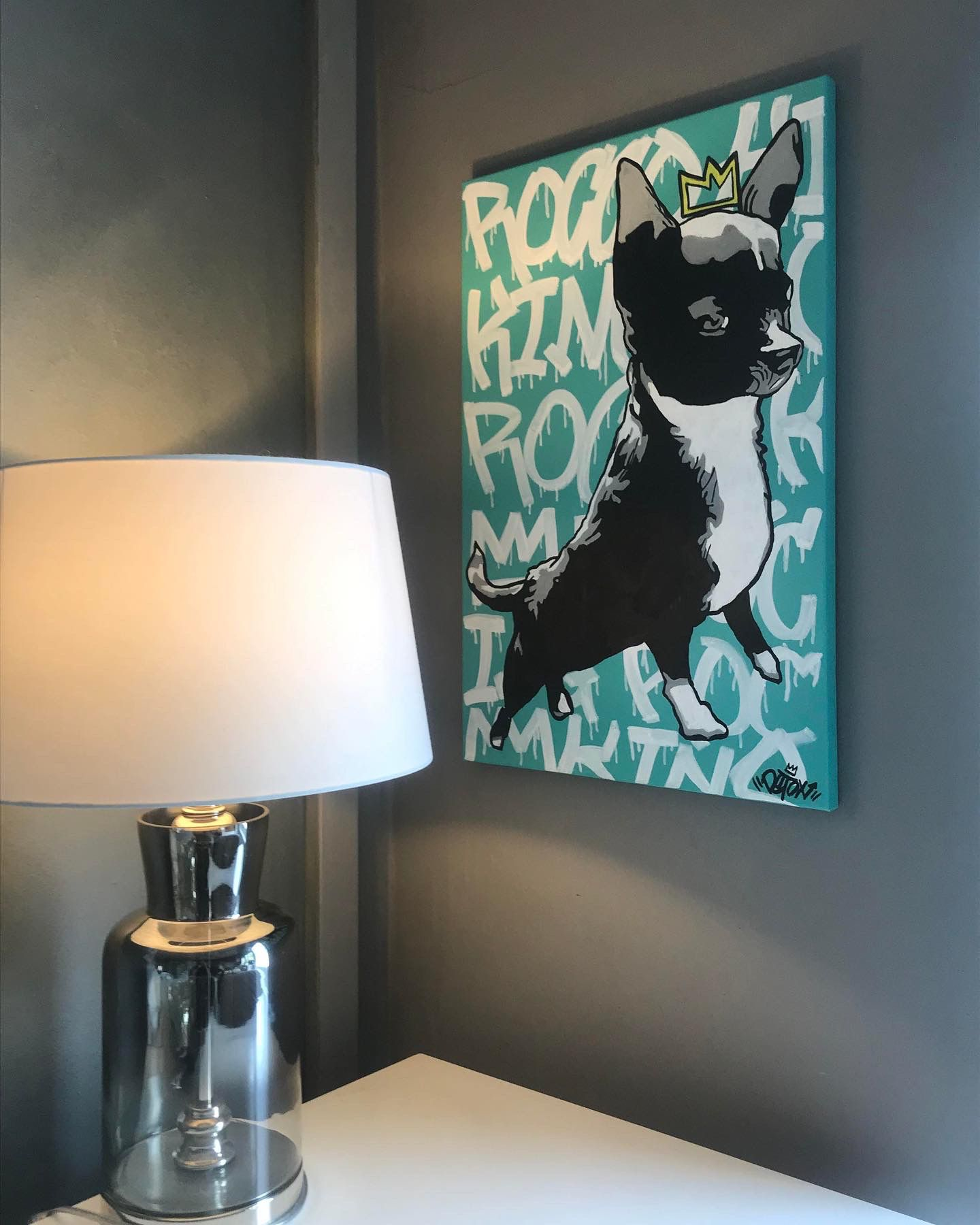 A modern living room corner with a silver lamp on a white table and a colorful painting of a French Bulldog with a crown on its head on the gray wall.