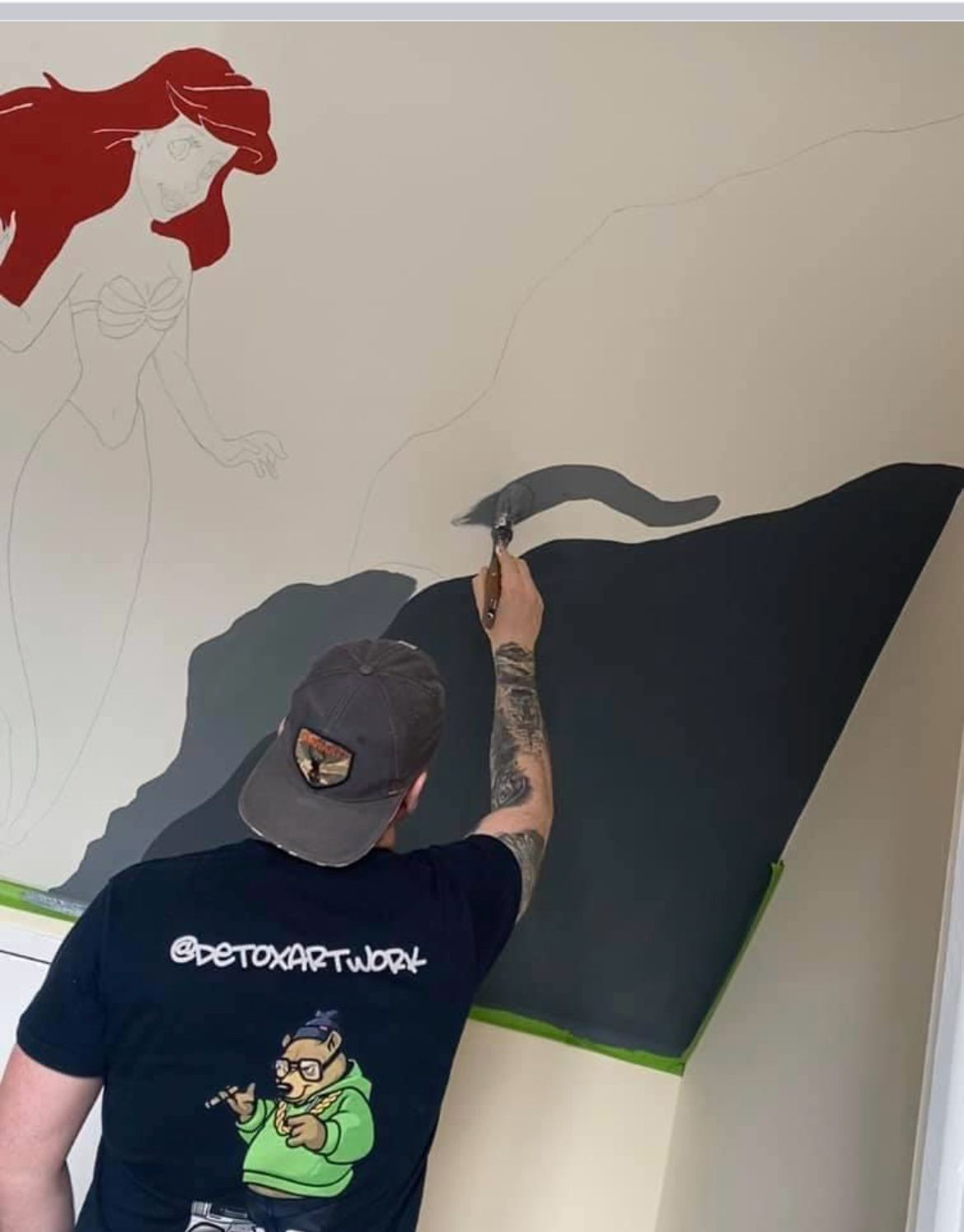 Artist painting a large mural of Ariel, the mermaid, on a ceiling with some parts still unfinished, including her tail and hair.